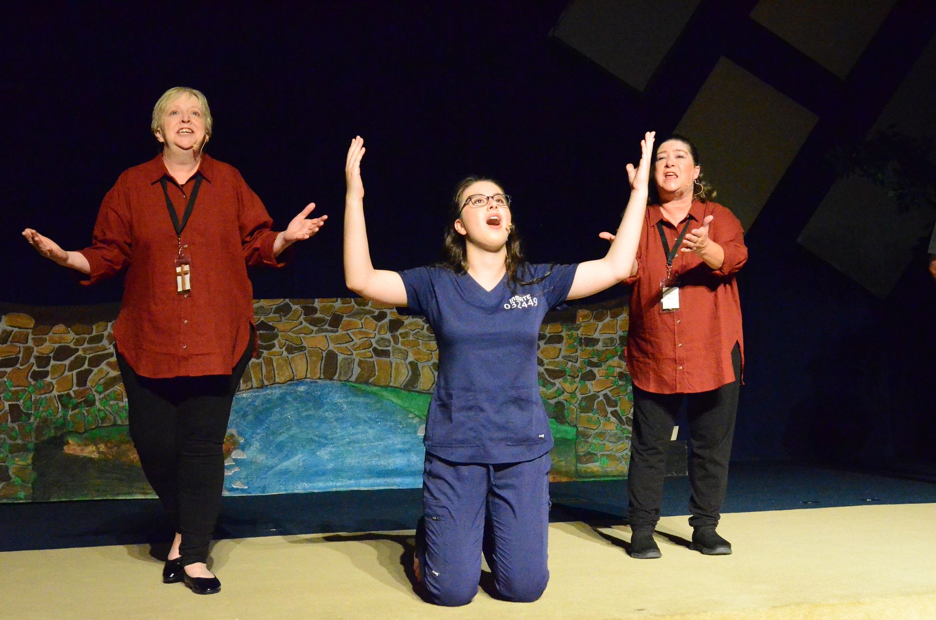 Three performers stand on stage against a backdrop depicting a stone wall and water, with two wearing red shirts and one wearing a blue shirt kneeling in the center with arms raised.