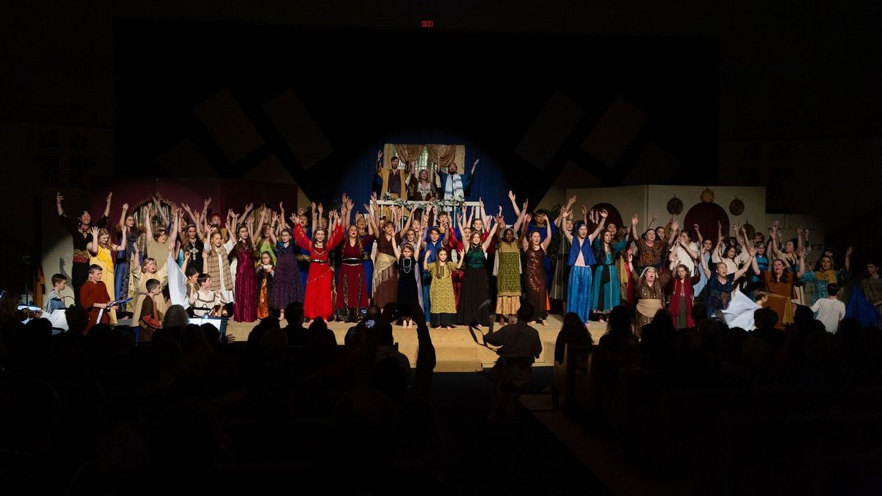 A large cast of performers in colorful period costumes stands on stage with arms raised in celebration, with a illuminated structure visible above them and audience members silhouetted in the foreground.