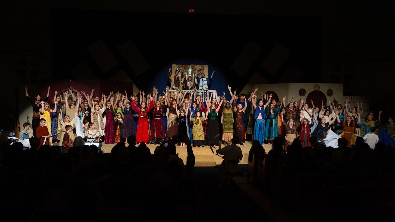 A large cast of performers in colorful period costumes stands on stage with arms raised in celebration, with a illuminated structure visible above them and audience members silhouetted in the foreground.