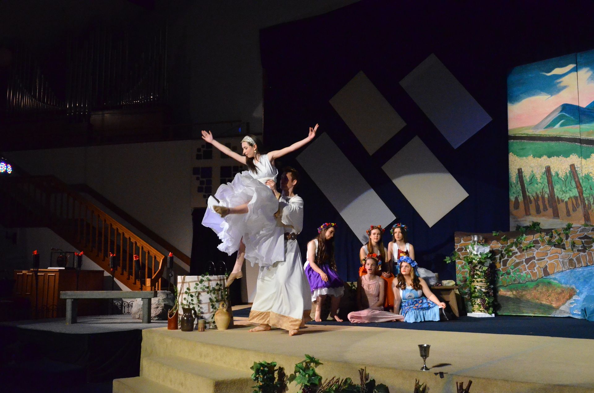 A performer in a white flowing costume stands with arms raised on stage surrounded by other cast members in period dress, with painted backdrop scenery of mountains and landscape visible behind them.