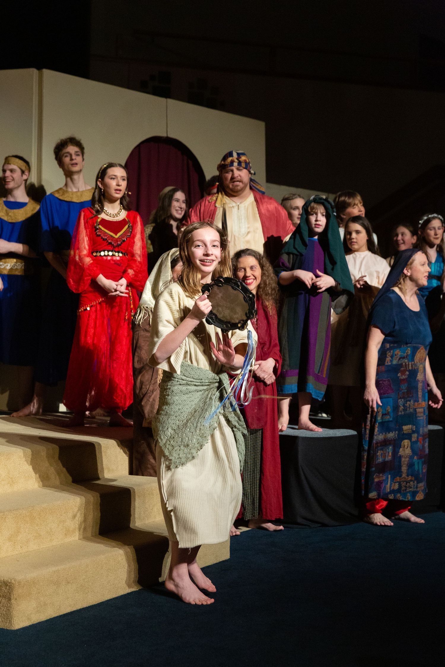 A diverse ensemble of performers in colorful period costumes stands together on a stepped stage set, with one performer in the foreground holding a tambourine and smiling toward the audience.