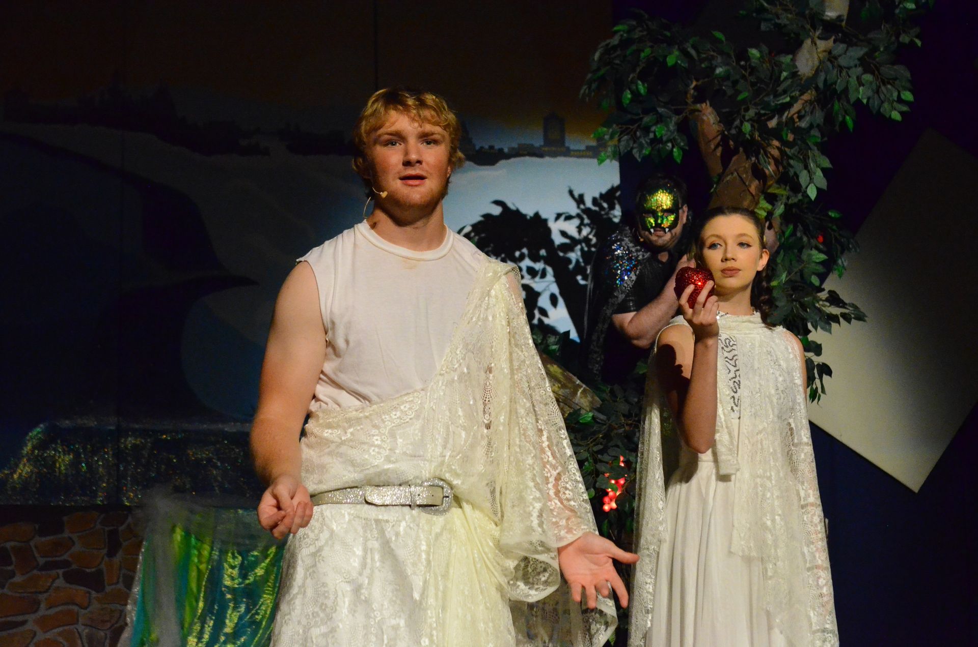 Two performers in flowing white and cream-colored robes stand center stage beside ivy-covered scenery, with one holding a red apple while a figure in elaborate green and gold costume emerges from the shadowed background.