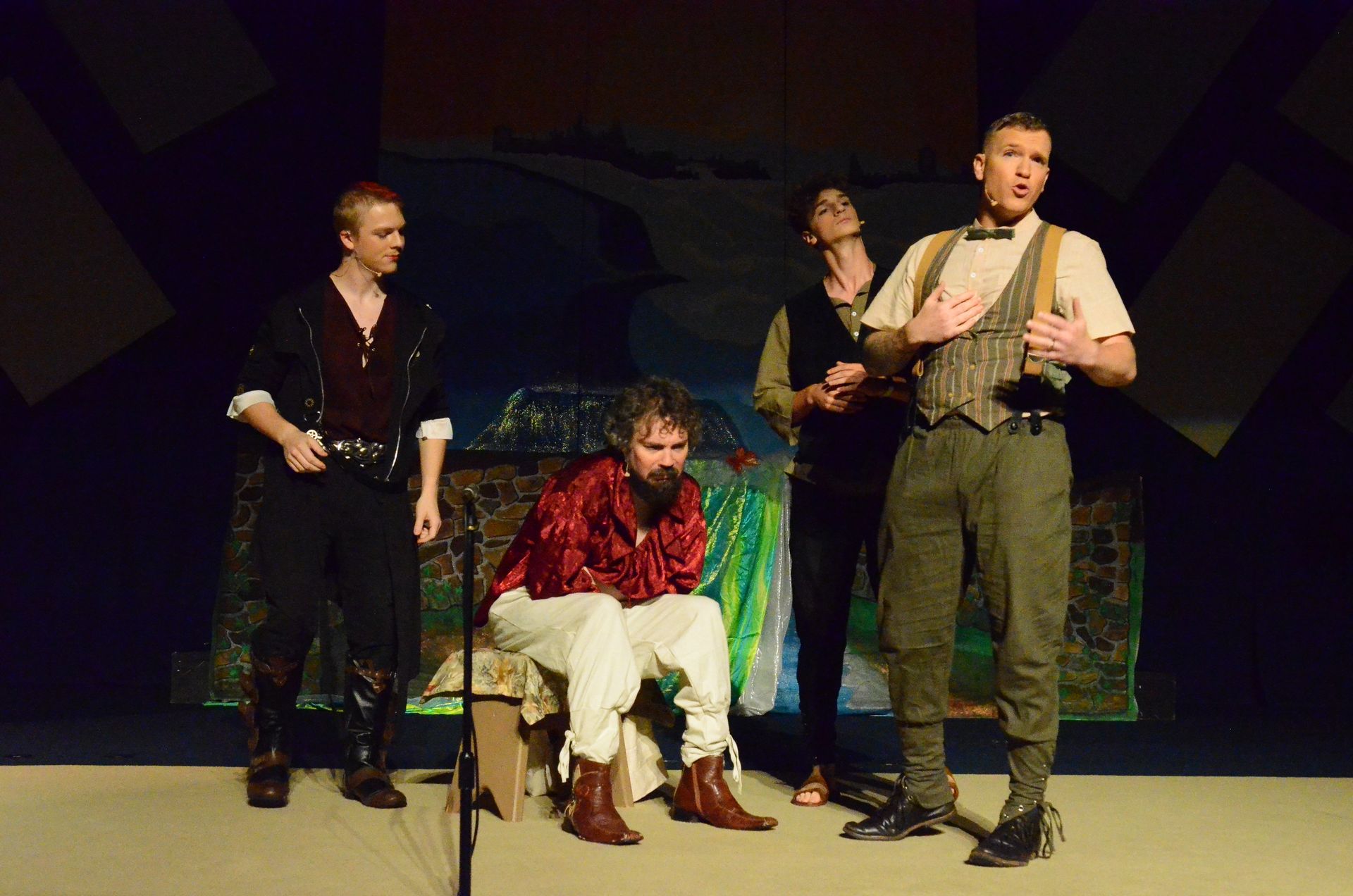 Four performers in period costumes stand on a stage with a painted backdrop depicting a biblical landscape, with one performer seated center stage wearing a red patterned robe and cream-colored pants while others stand around in various earth-toned garments.