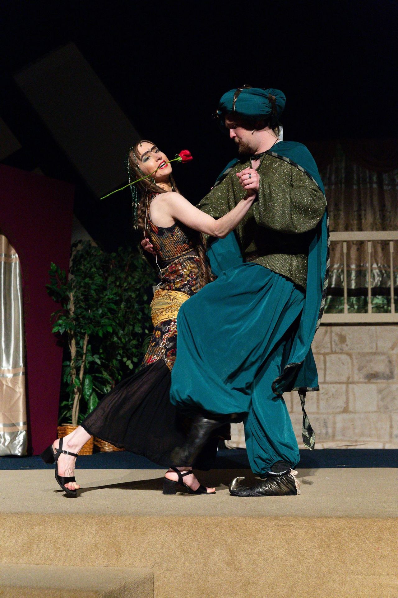 Two performers in medieval-style costumes dance together on stage, with one holding a red rose, set against a backdrop featuring a stone building and burgundy wall.