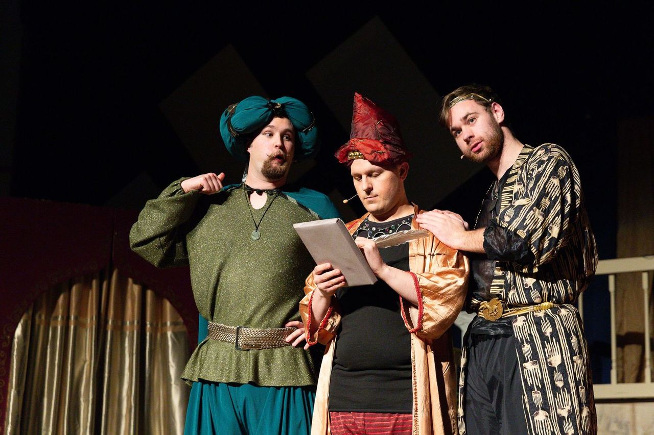 Four performers in medieval-style costumes stand together on stage, with one holding a tablet, against a dark backdrop with geometric shapes.
