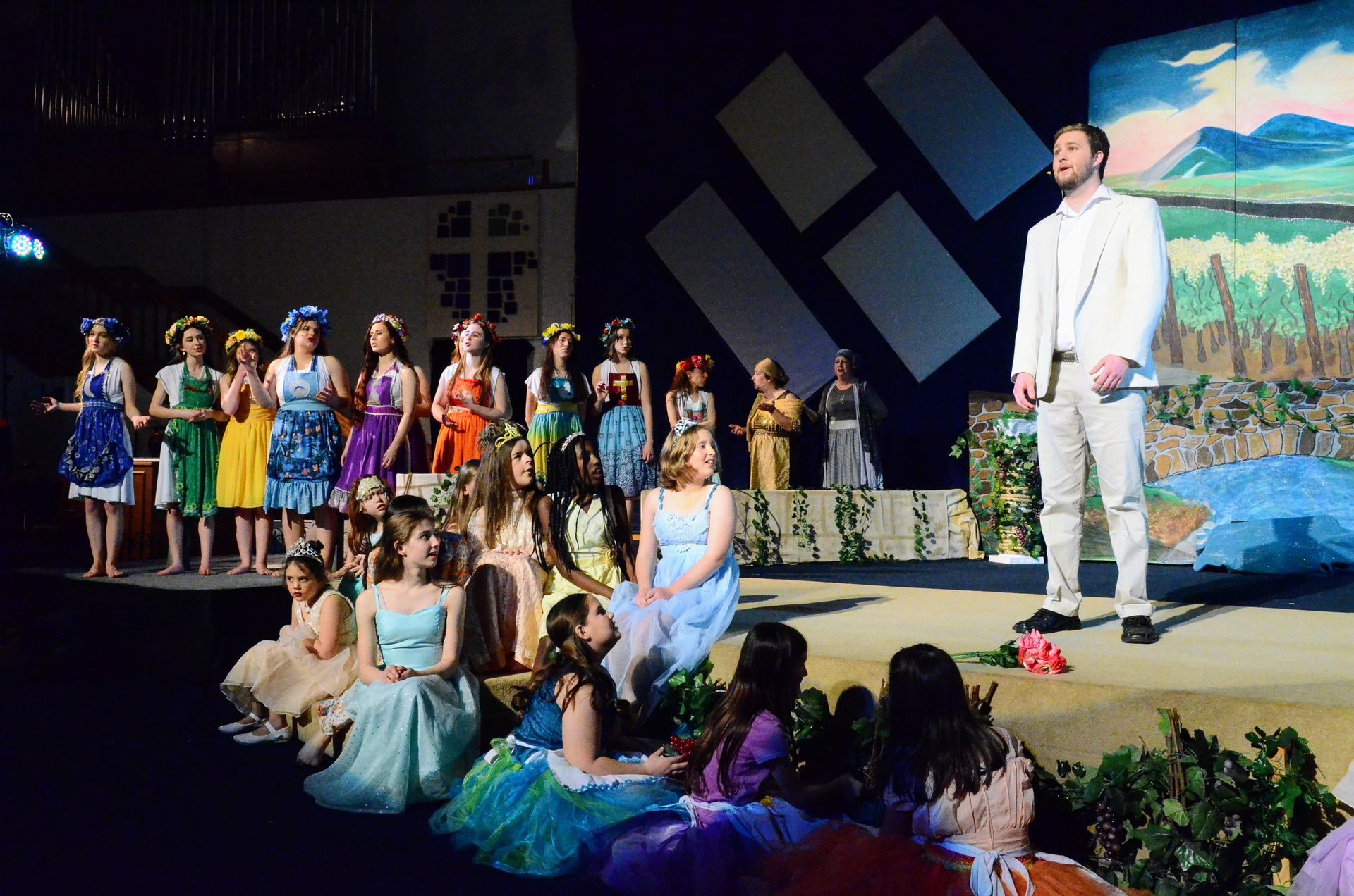 A cast of performers in colorful period costumes stands and sits on stage surrounded by potted plants, with a male performer in a cream-colored suit standing to the right, while painted backdrops of pastoral landscapes and geometric shapes create the theatrical setting.
