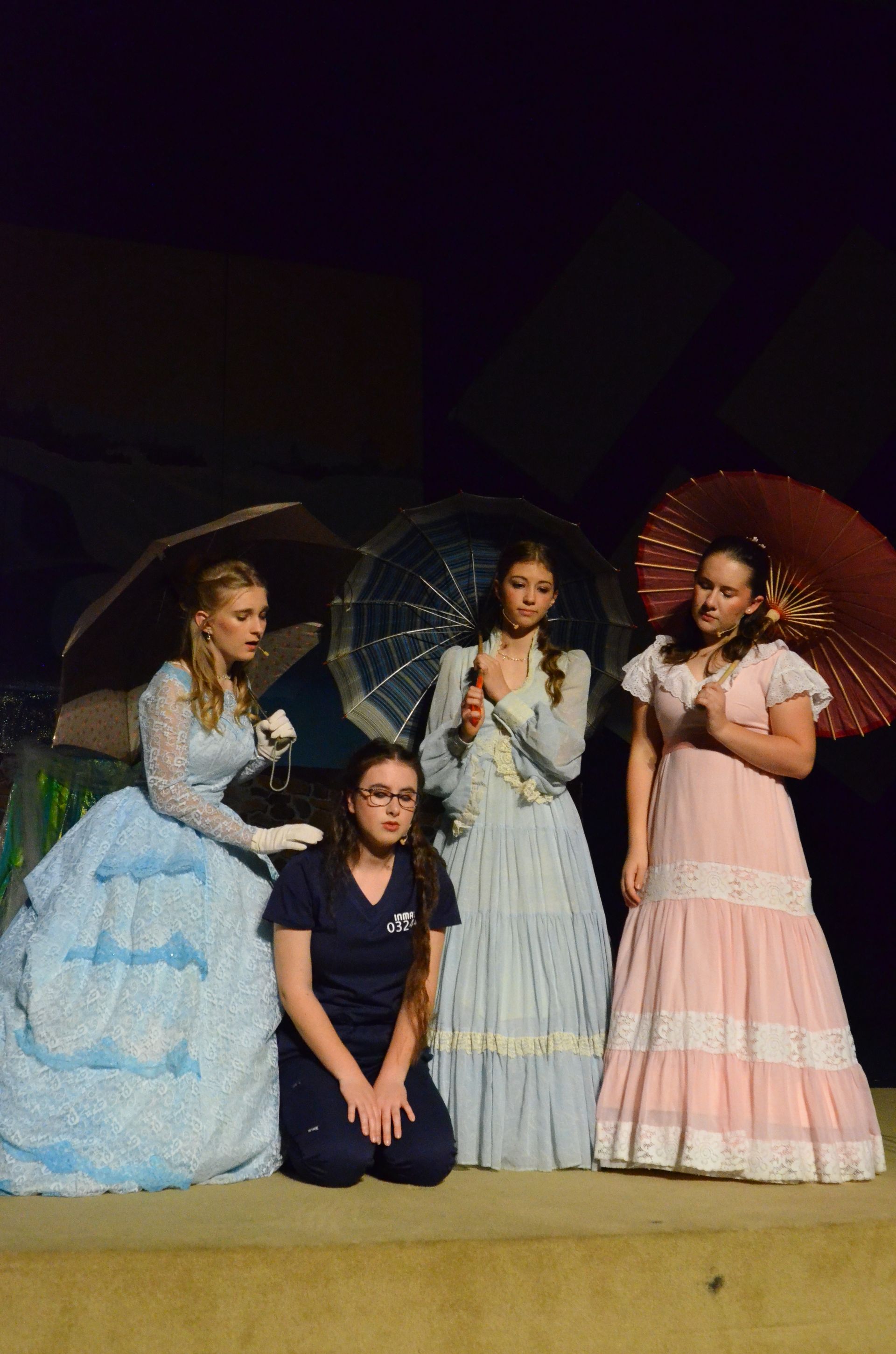 Four performers in period costumes stand against a yellow wall, with two holding decorative parasols, dressed in pastel-colored gowns and casual clothing in a theatrical scene lit against a dark background.