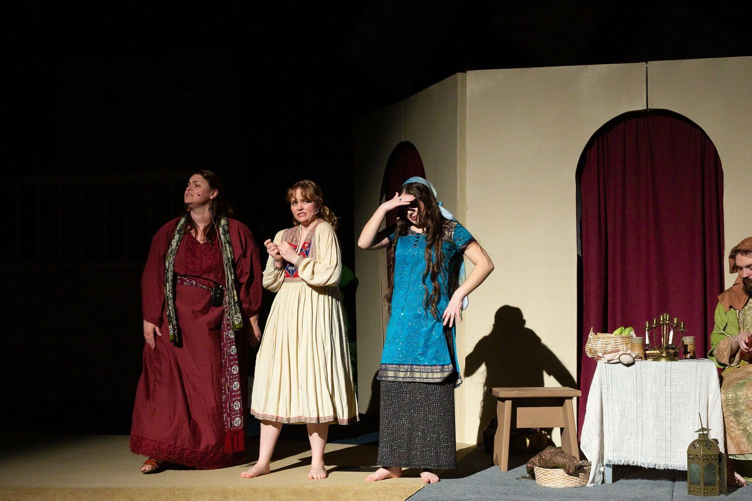 Four performers in period costumes stand on a stage set with beige walls, a burgundy curtained doorway, and a table with props, while one performer sits at a wooden chair in the scene.