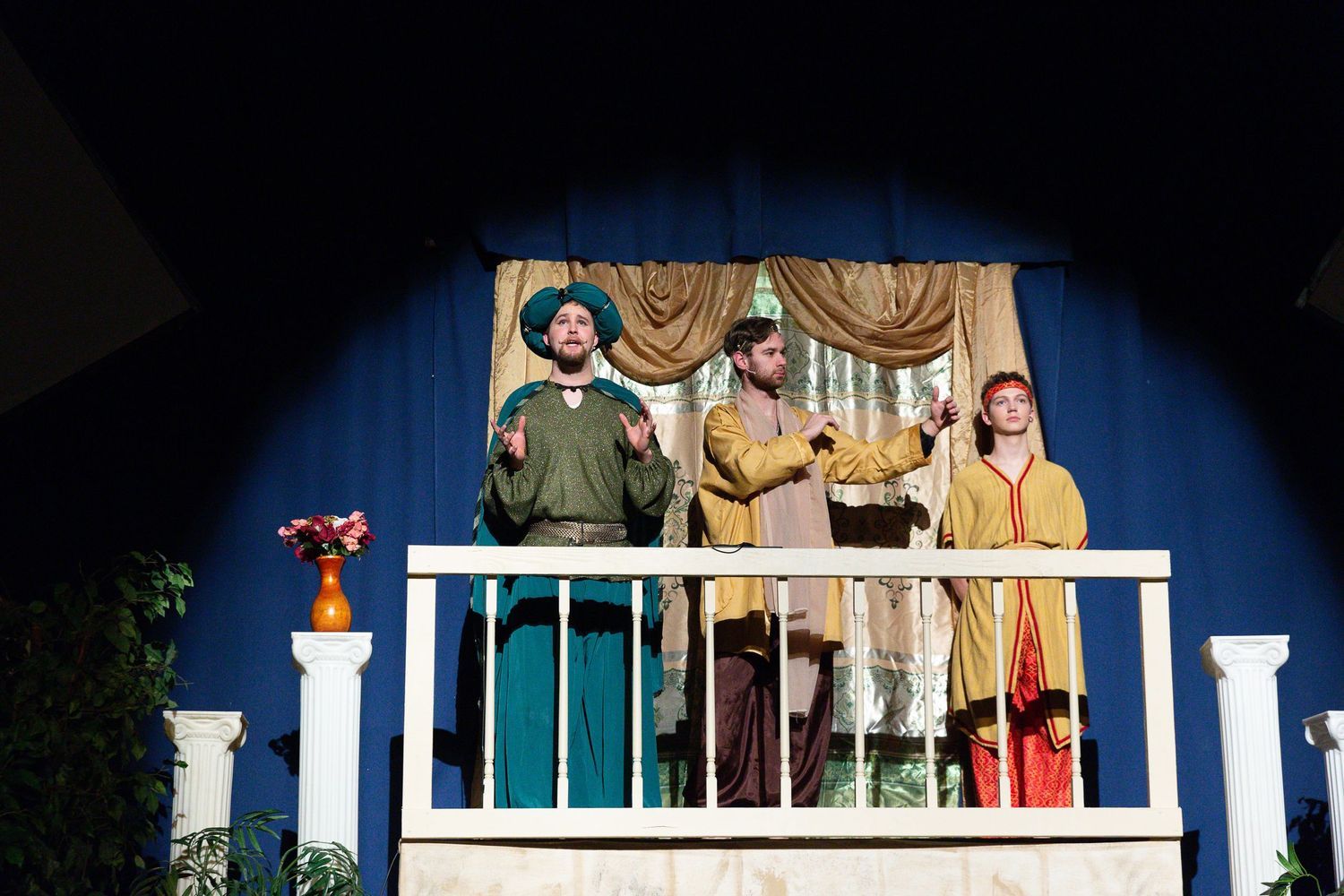 Three performers in medieval-style costumes stand on a white-railed balcony stage set against a blue backdrop with gold draped curtains, potted flowers at the railings, and classical white columns flanking the scene.