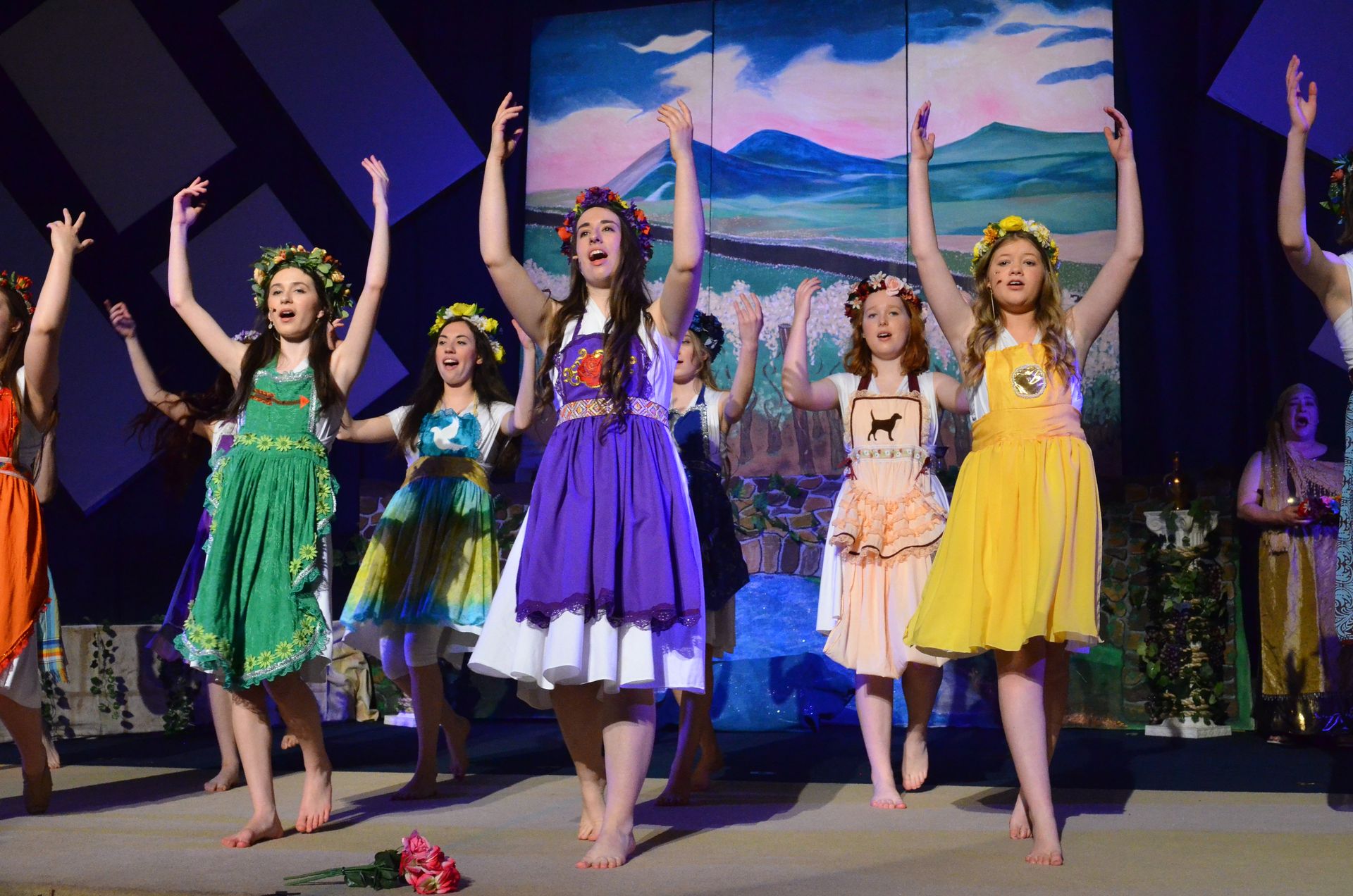 A cast of performers wearing colorful floral-adorned dresses and flower crowns performs on stage with their arms raised in celebration, with a scenic painted backdrop of mountains and landscape behind them.