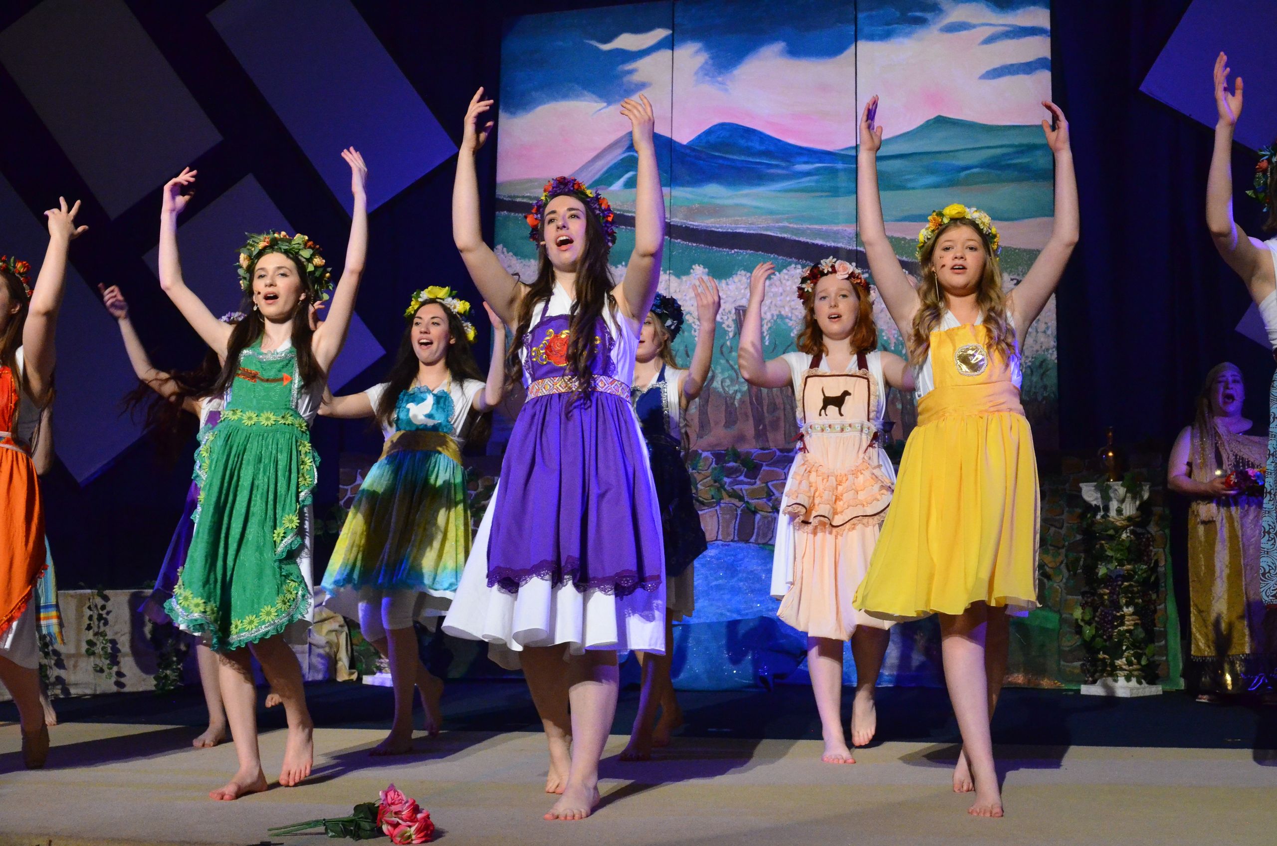 A cast of performers wearing colorful floral-adorned dresses and flower crowns performs on stage with their arms raised in celebration, with a scenic painted backdrop of mountains and landscape behind them.