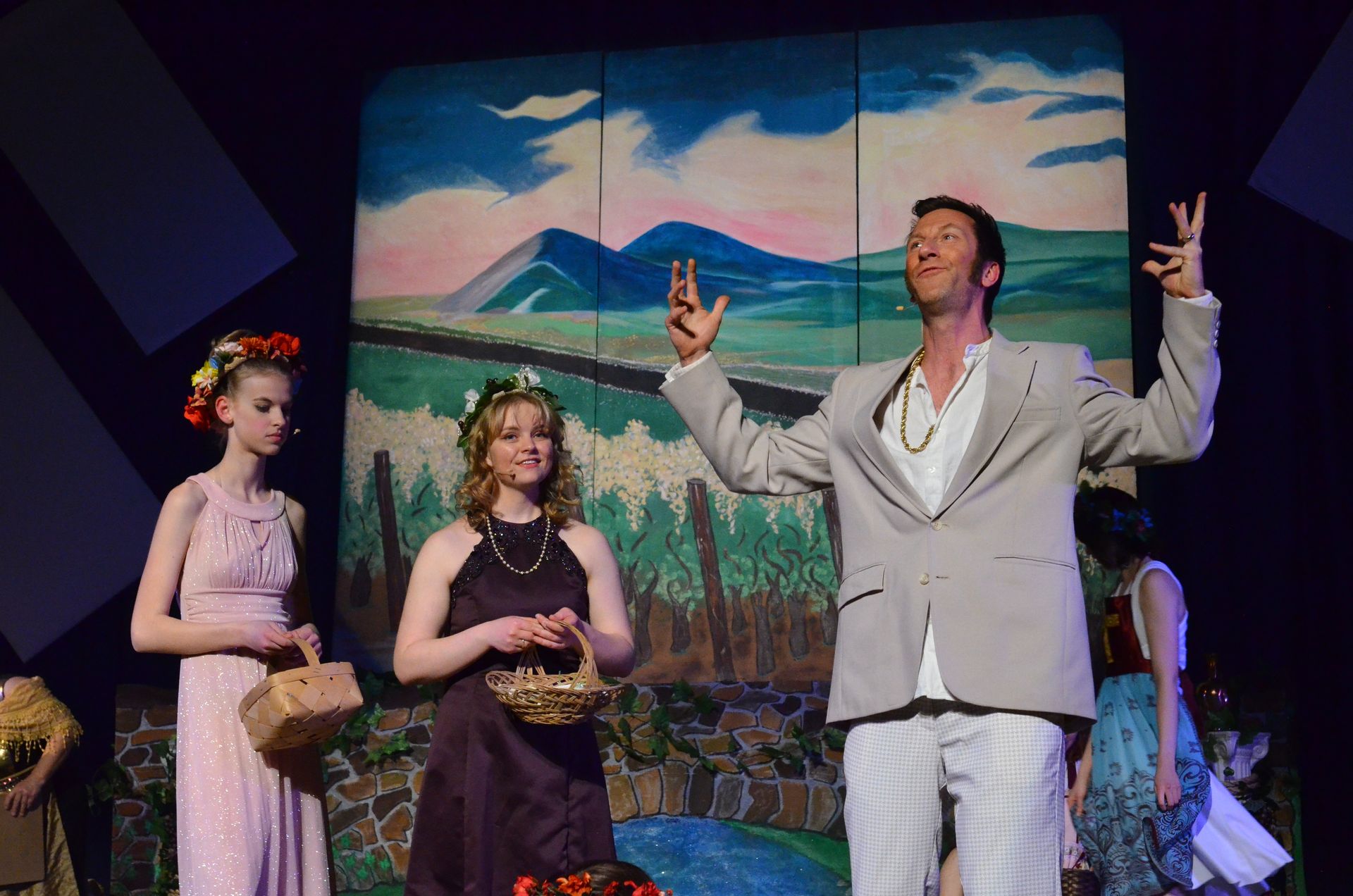 A performer in a cream-colored suit gestures expressively toward the sky while two performers in floral crowns hold baskets beside him, all positioned in front of a painted backdrop depicting mountains, fields, and a pastoral landscape.