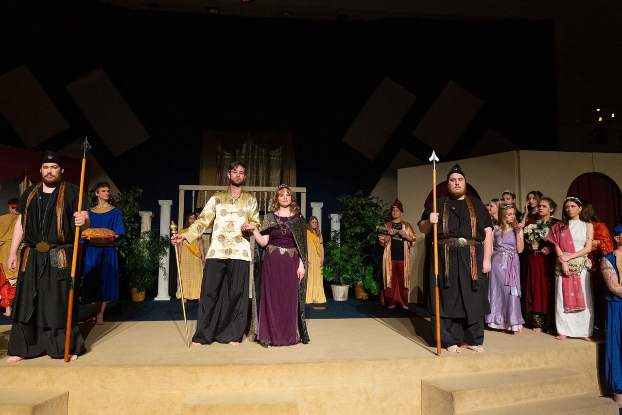 A large cast of performers in medieval-style robes and tunics stands on a sand-colored stage set with classical columns and greenery, with two performers holding tall staffs at center stage and ensemble members arranged on both sides.