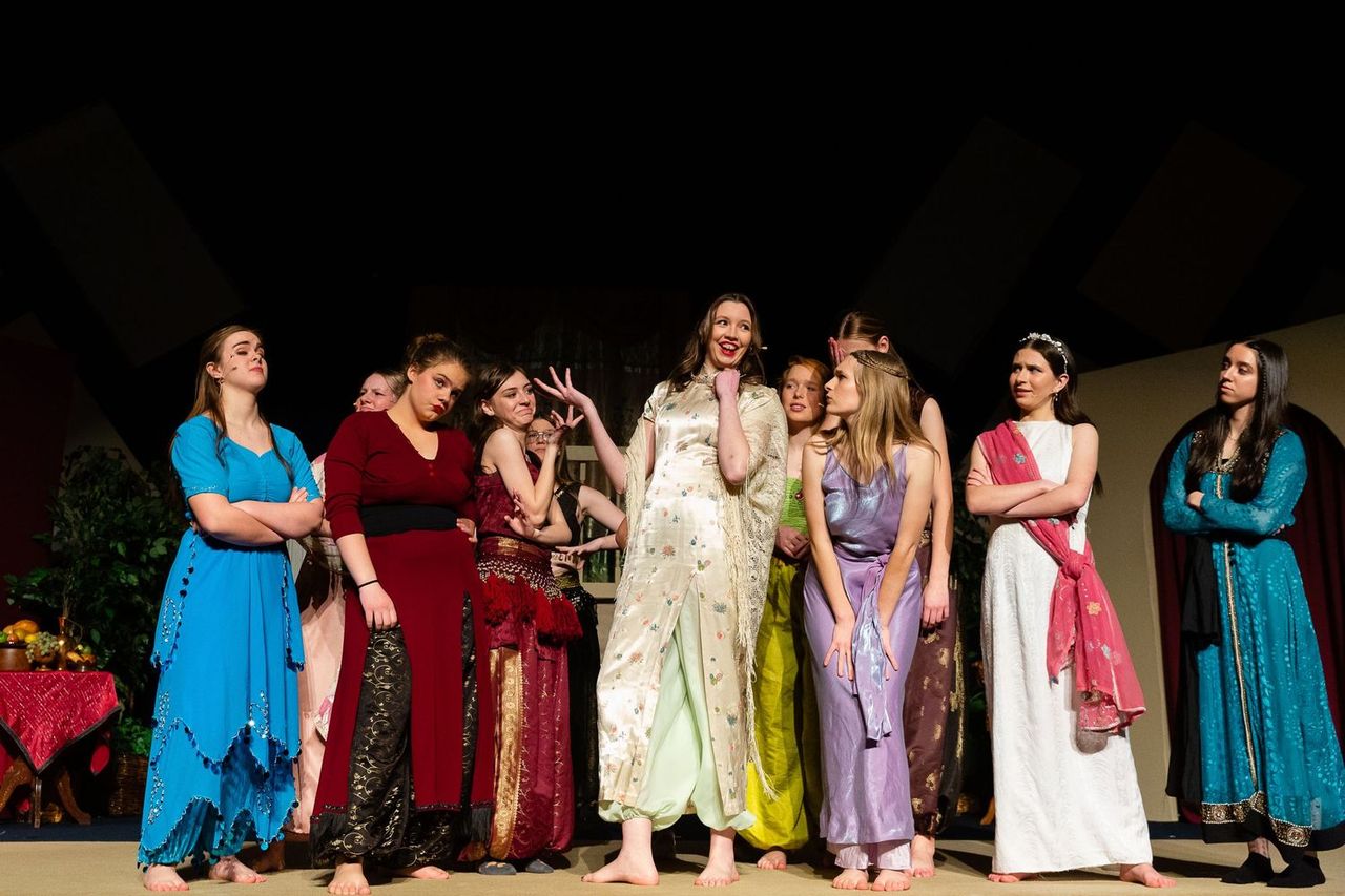 A cast of performers in colorful period costumes stands together on stage under theatrical lighting, with one performer in a cream-colored dress with floral patterns standing prominently in the center while others gesture expressively around her.