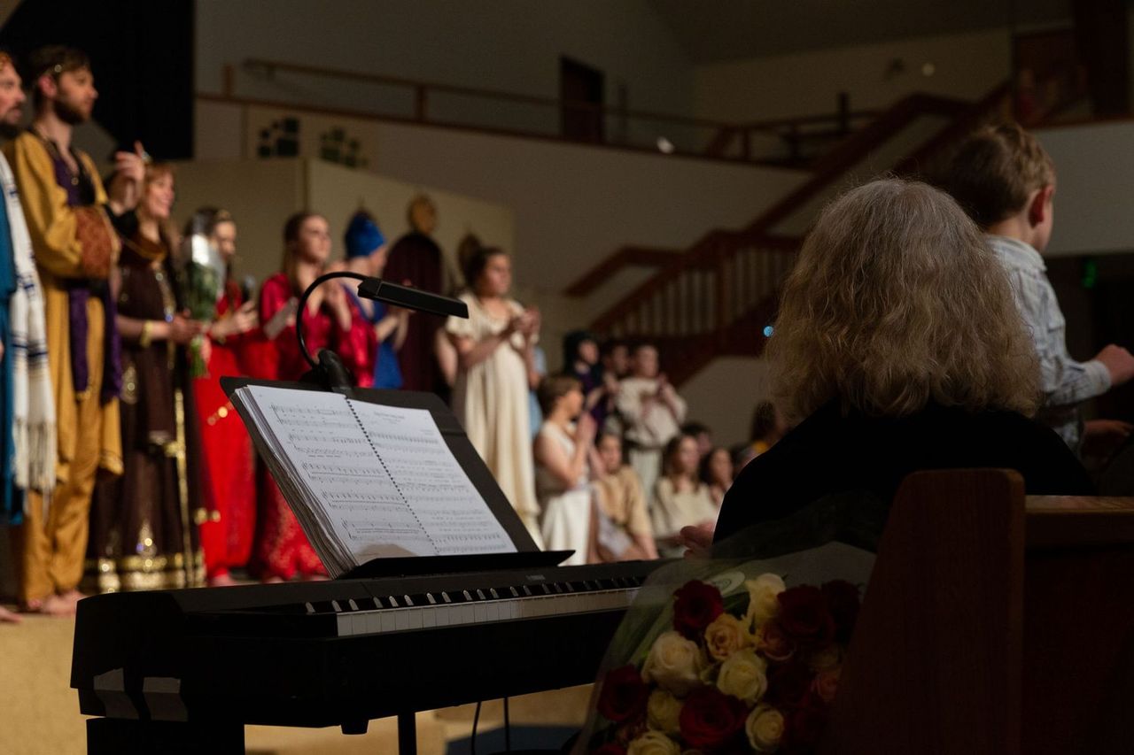A pianist with gray hair performs at a keyboard while reading sheet music, accompanying a cast of performers in colorful period costumes singing on stage in a gymnasium setting.