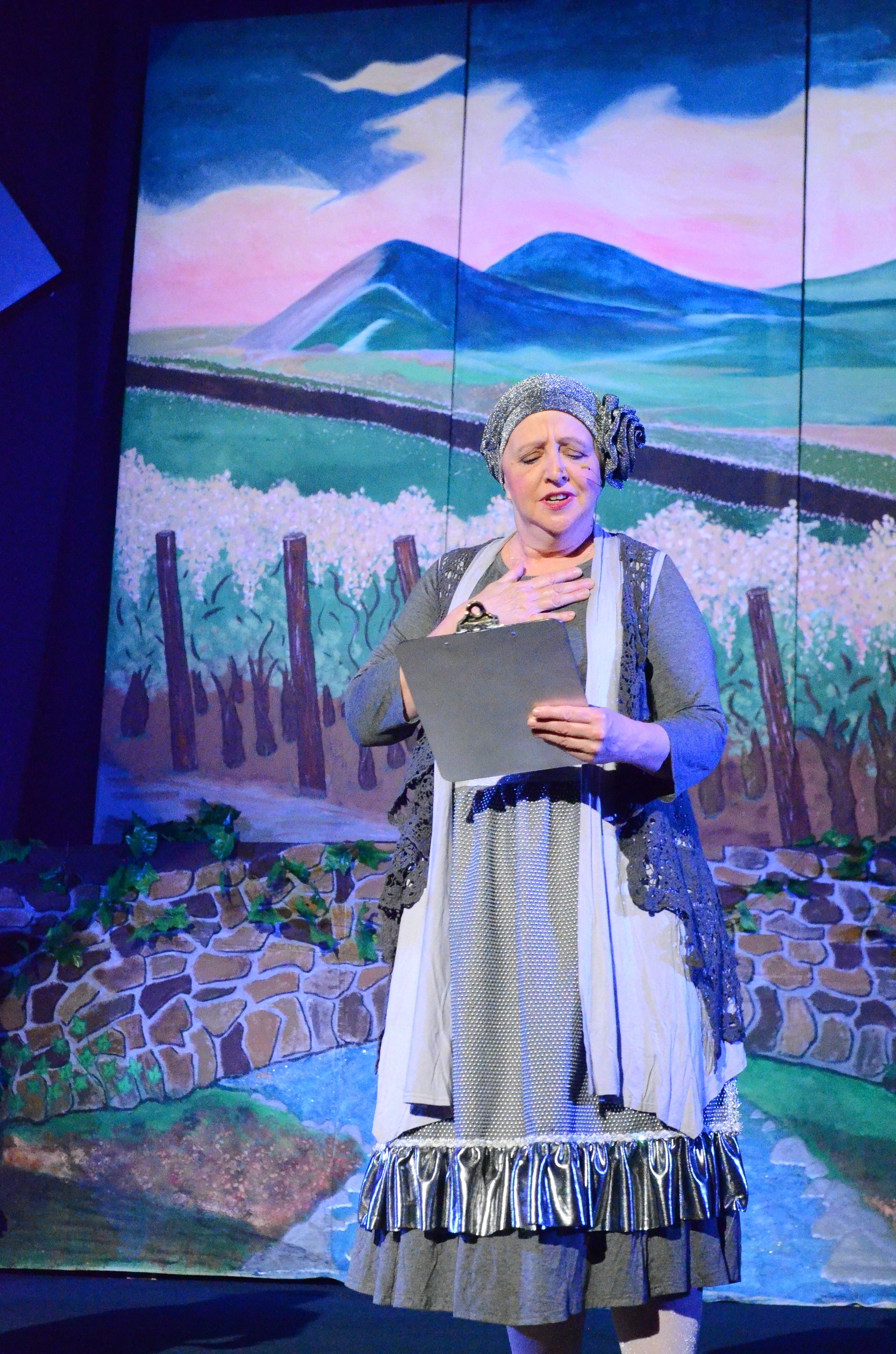 A performer in a striped dress and grey shawl stands center stage against a colorful projected backdrop of blue, green, and pink hues depicting an underwater or mystical scene.
