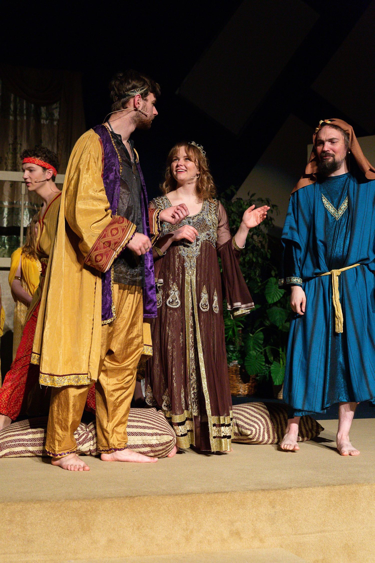 Four performers in colorful medieval-style robes and tunics stand on a sandy stage with striped floor coverings and green foliage props, engaging in conversation during a theatrical scene.