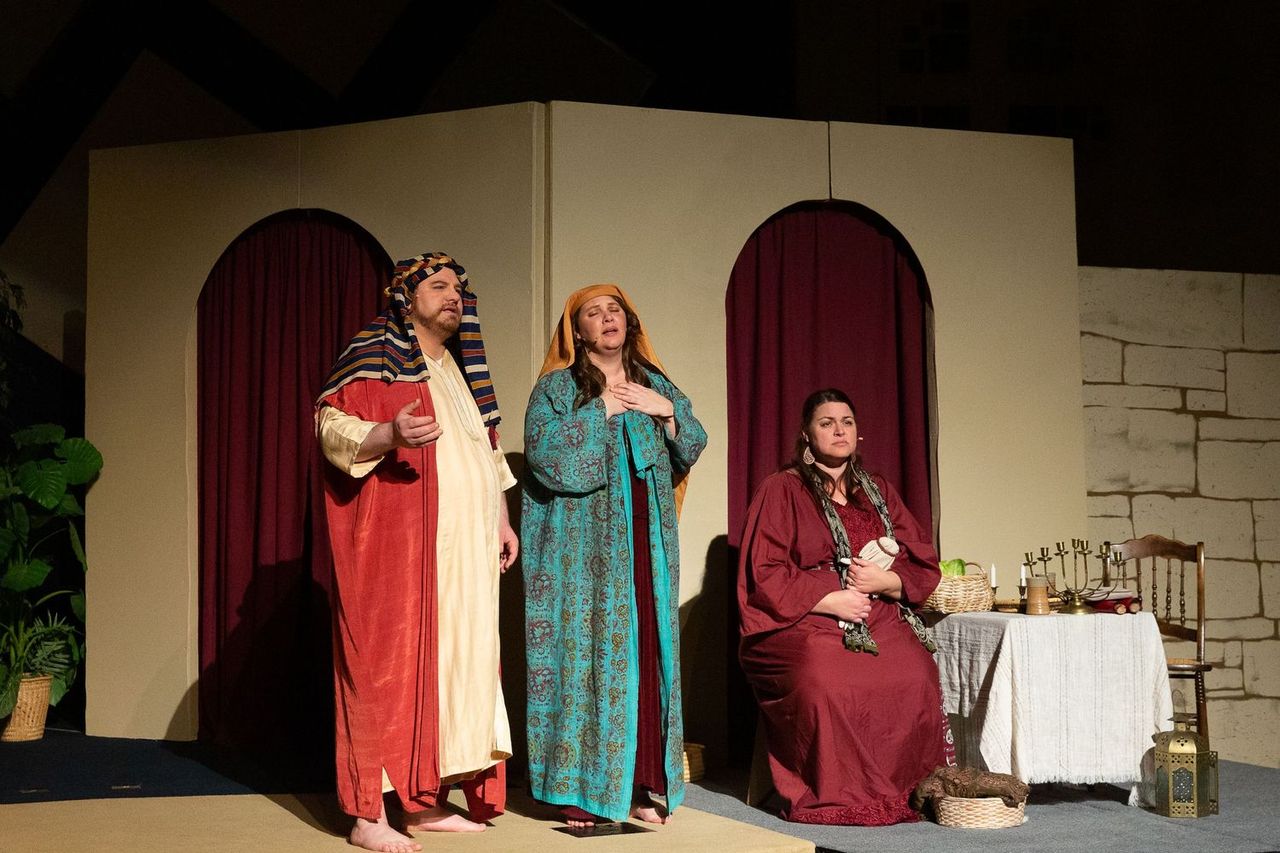 Three performers in elaborate biblical-era costumes stand on a stage set with arched doorways, red curtains, and period furnishings, with two standing center stage and one seated at a table with props.