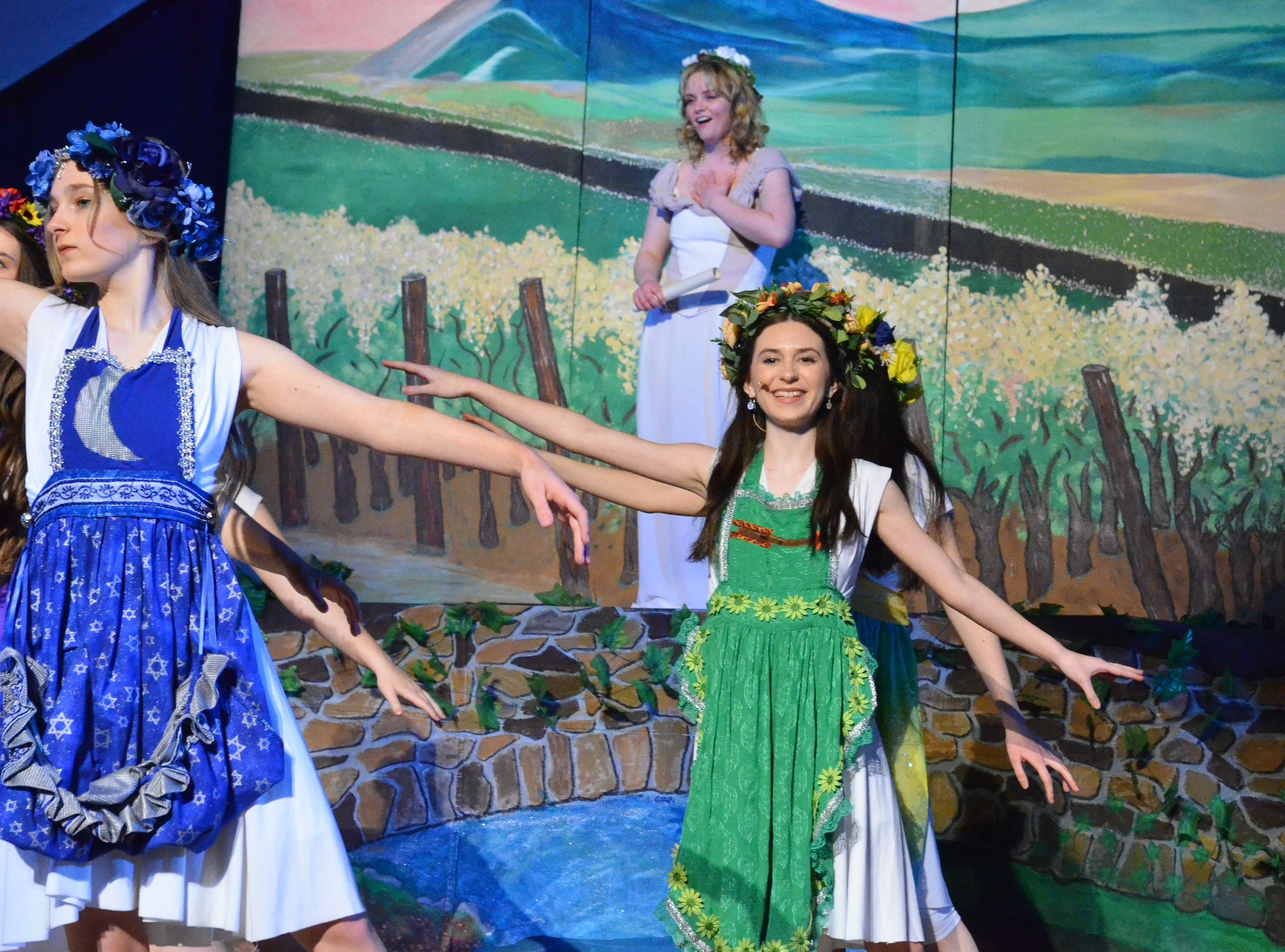 Two performers in colorful folk-style costumes with floral crowns dance expressively on stage before a painted backdrop depicting a rural landscape with fields, wooden fences, and a distant horizon.