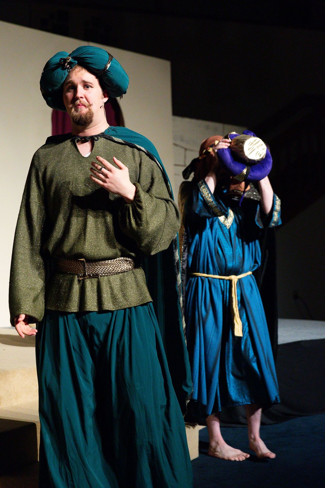 Two performers on stage wear teal and blue tunics with matching head coverings, with one standing at center stage with hand to chest while the other stands behind holding a lamp.