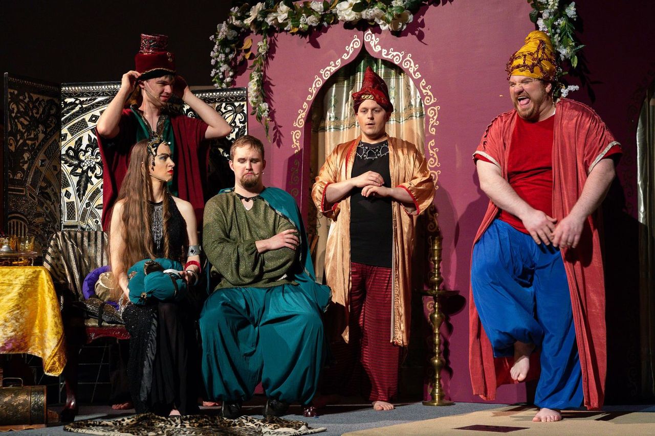 Five performers in elaborate period costumes stand on a theatrical stage with a magenta backdrop, ornate panels, floral garlands, and decorative furnishings, with two seated and three standing in various poses of engagement.