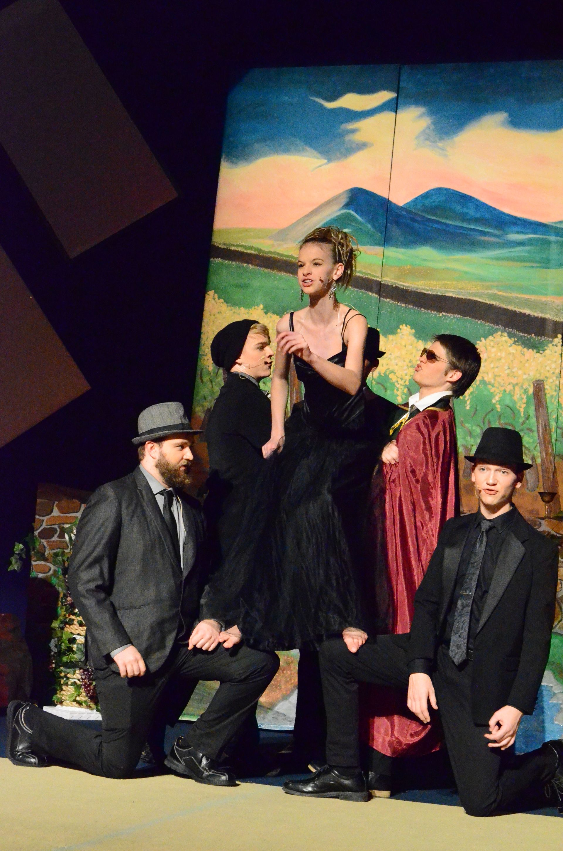 A cast of five performers in dark formal attire poses together against a vibrant painted backdrop featuring green, blue, and warm-toned abstract scenery.