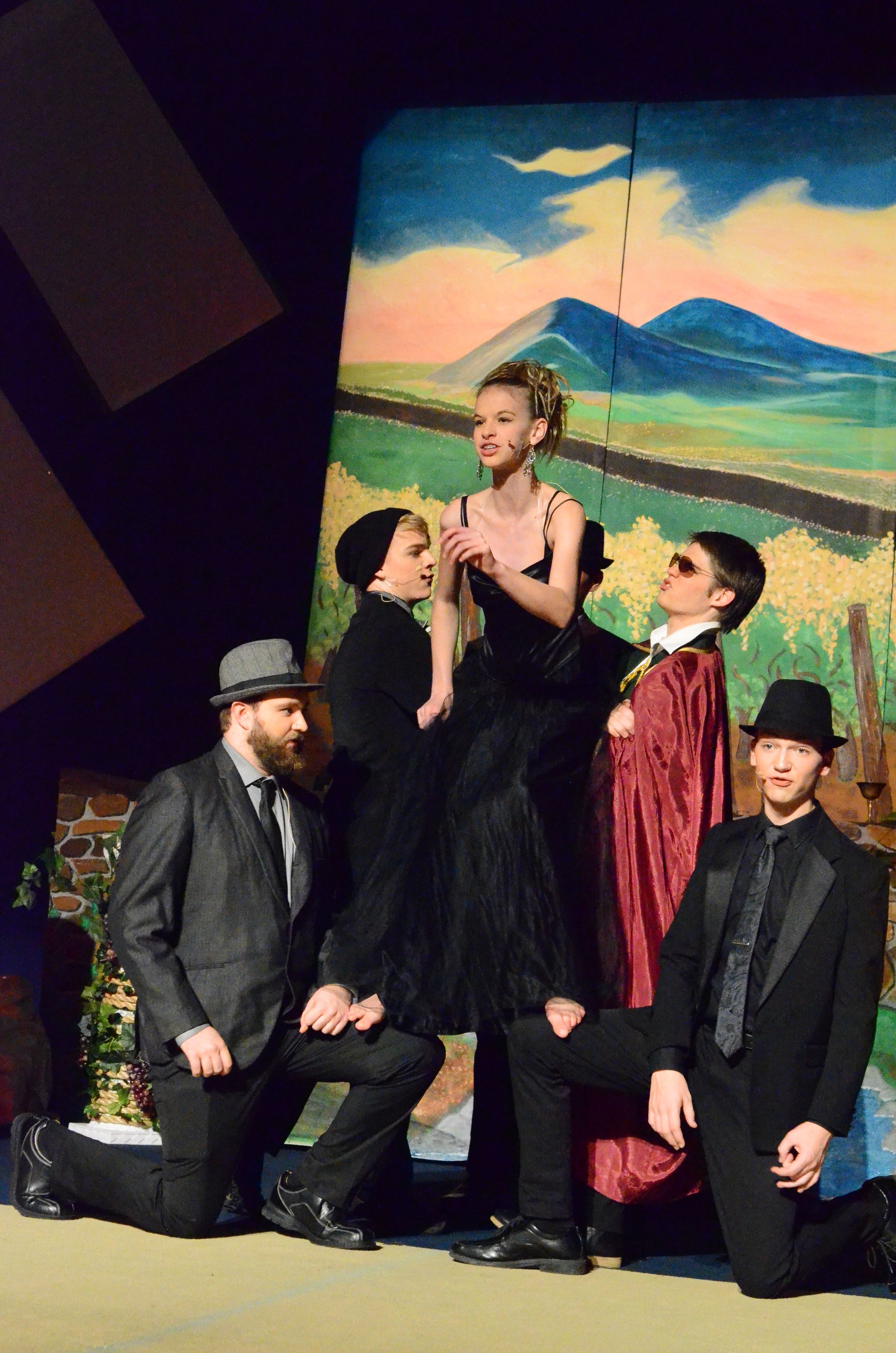 A cast of five performers in dark formal attire poses together against a vibrant painted backdrop featuring green, blue, and warm-toned abstract scenery.