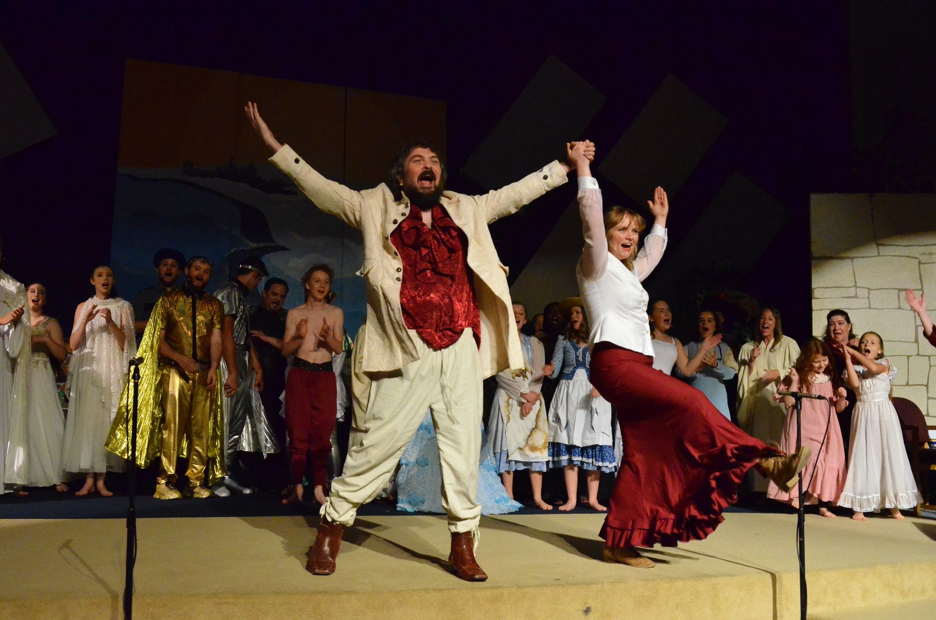 Two performers in colorful period costumes leap joyfully on stage with their arms raised while a large ensemble cast in flowing robes and tunics watches from behind them on an indoor theatre stage.