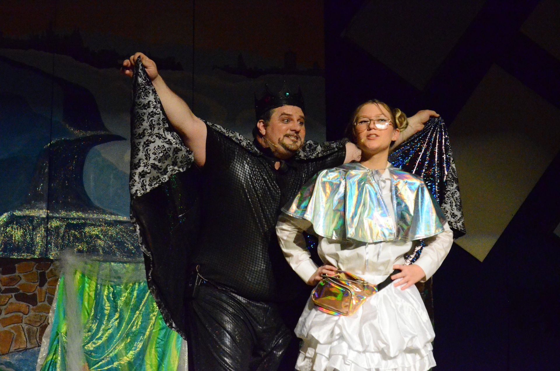 Two performers on stage wear elaborate costumes—one in a black mesh outfit with ornate lace sleeves and a cap, the other in a white ruffled dress with a holographic cape and glasses—as they hold up decorative fabric against a theatrical backdrop featuring green moss-covered scenery and stone textures.