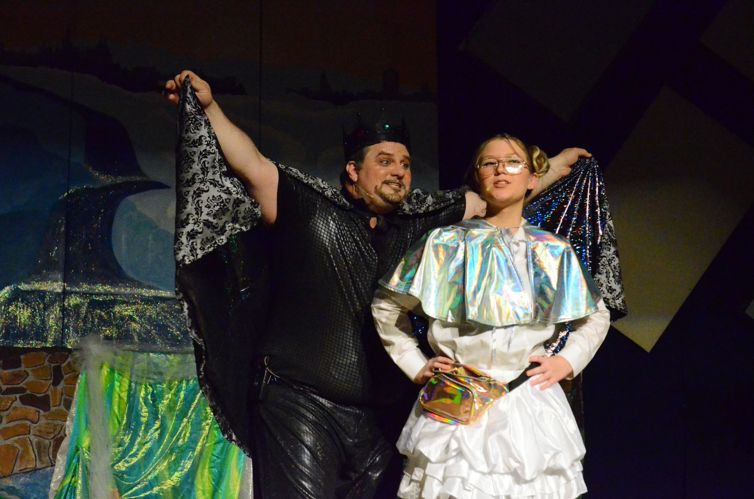 Two performers on stage wear elaborate costumes—one in a black mesh outfit with ornate lace sleeves and a cap, the other in a white ruffled dress with a holographic cape and glasses—as they hold up decorative fabric against a theatrical backdrop featuring green moss-covered scenery and stone textures.