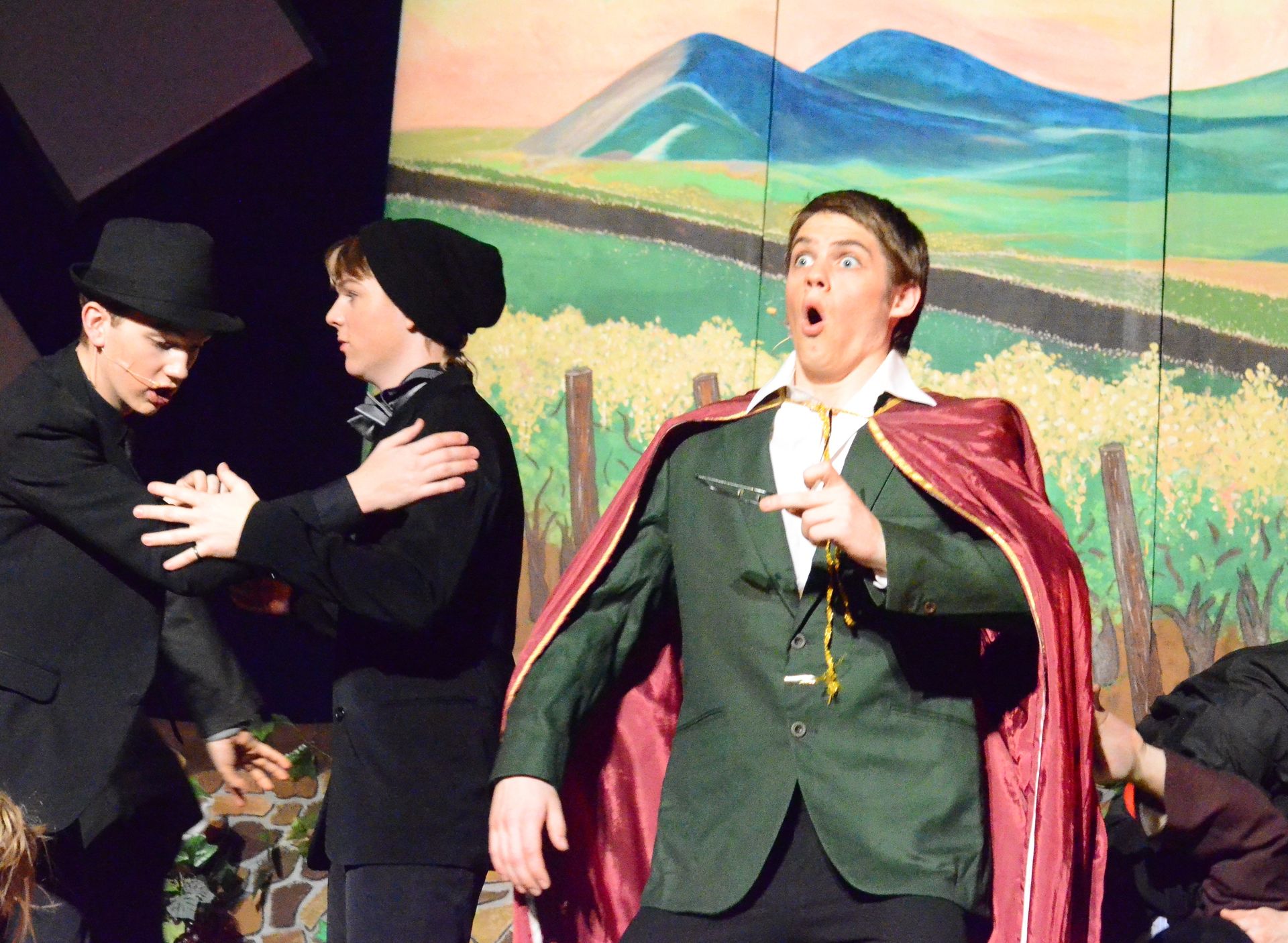 Three performers in period costumes stand on stage before a painted backdrop of rolling green fields and blue mountains, with two wearing black coats and hats on the left and one on the right wearing a green jacket with a dramatic red cape while gesturing expressively.