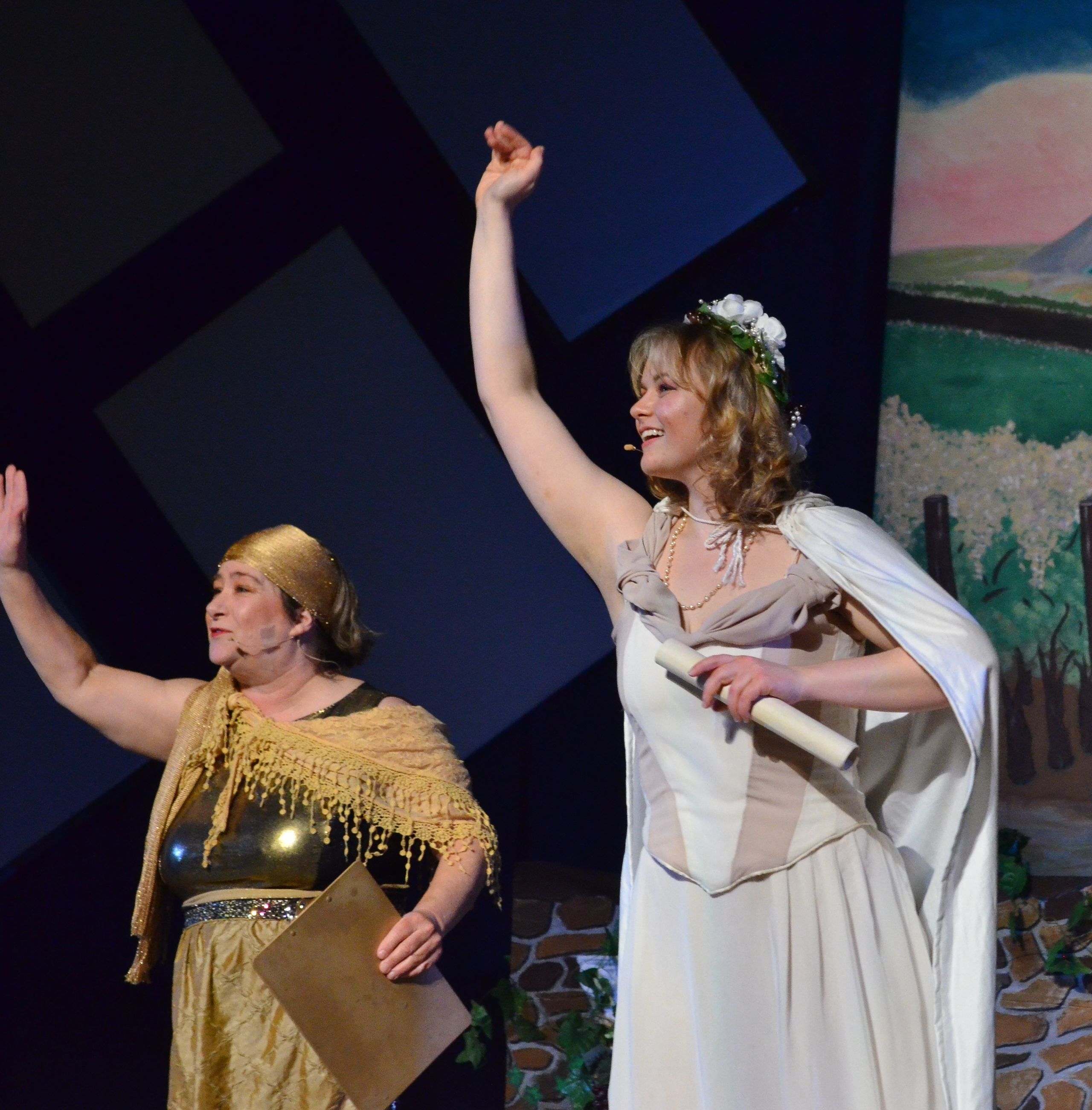 Two performers in classical robes stand on stage with their arms raised triumphantly, one wearing a gold draped cape and the other in a white gown with a flower crown and white shawl, against a stylized backdrop of geometric shapes and landscape scenery.