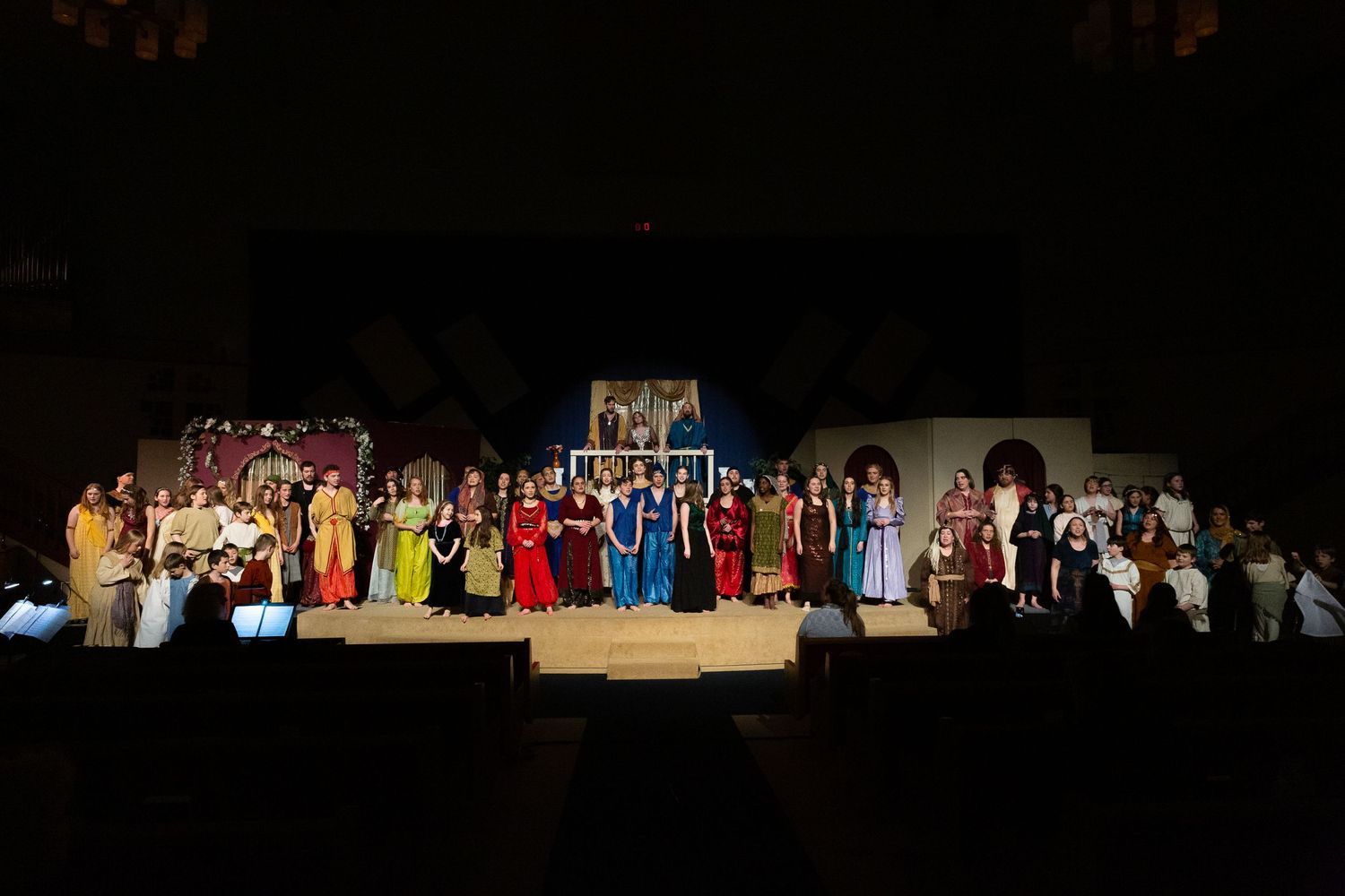 A large cast of performers in colorful period costumes stands together on a brightly lit stage with ornate set pieces and a painted backdrop, taking their final bow.