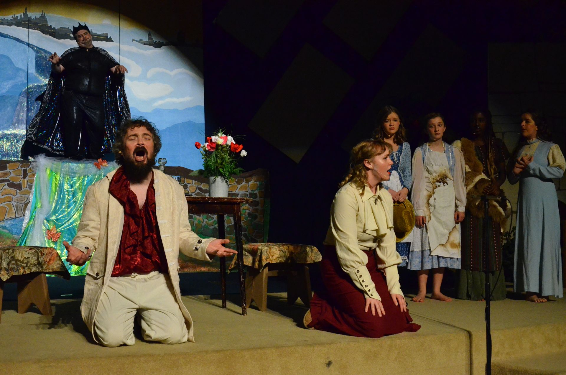 A male performer kneels on stage in a cream-colored suit and burgundy shirt with an expressive face, while a female performer in similar period costume kneels nearby, with additional cast members standing in the background on a set featuring a projected backdrop of a coastal landscape.