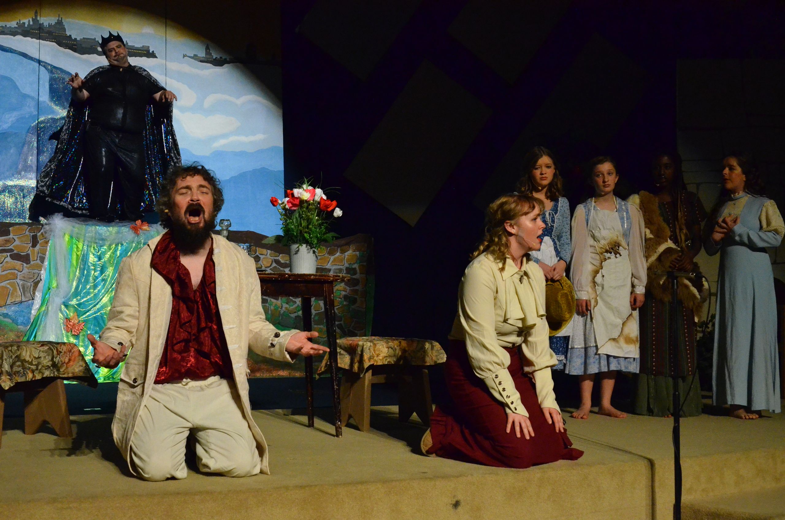 A male performer kneels on stage in a cream-colored suit and burgundy shirt with an expressive face, while a female performer in similar period costume kneels nearby, with additional cast members standing in the background on a set featuring a projected backdrop of a coastal landscape.