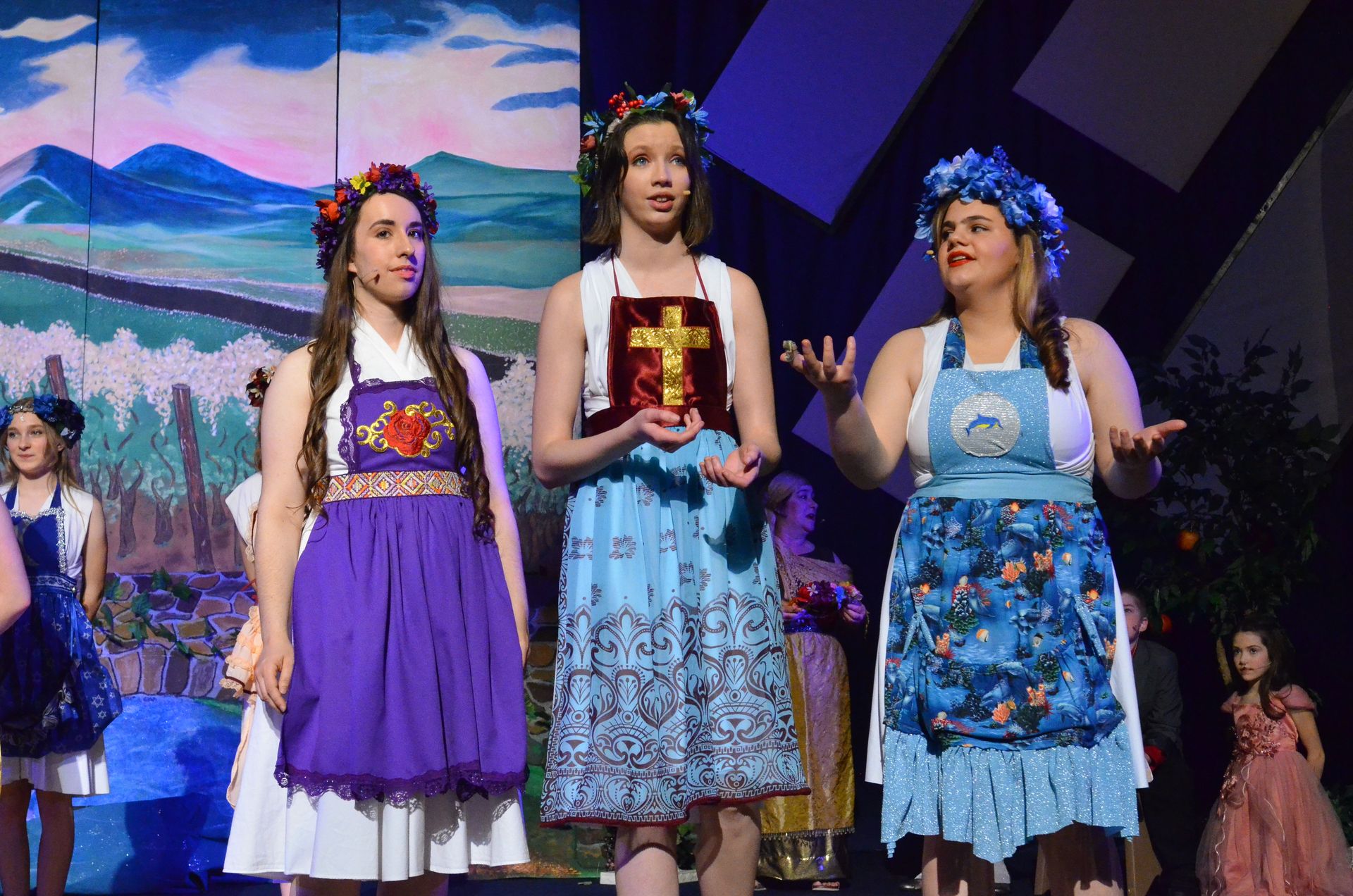 Three performers wearing colorful floral crowns and traditional-style dresses in shades of purple, white, and blue stand on stage before a painted backdrop depicting mountains and a landscape, with additional cast members visible in the background.