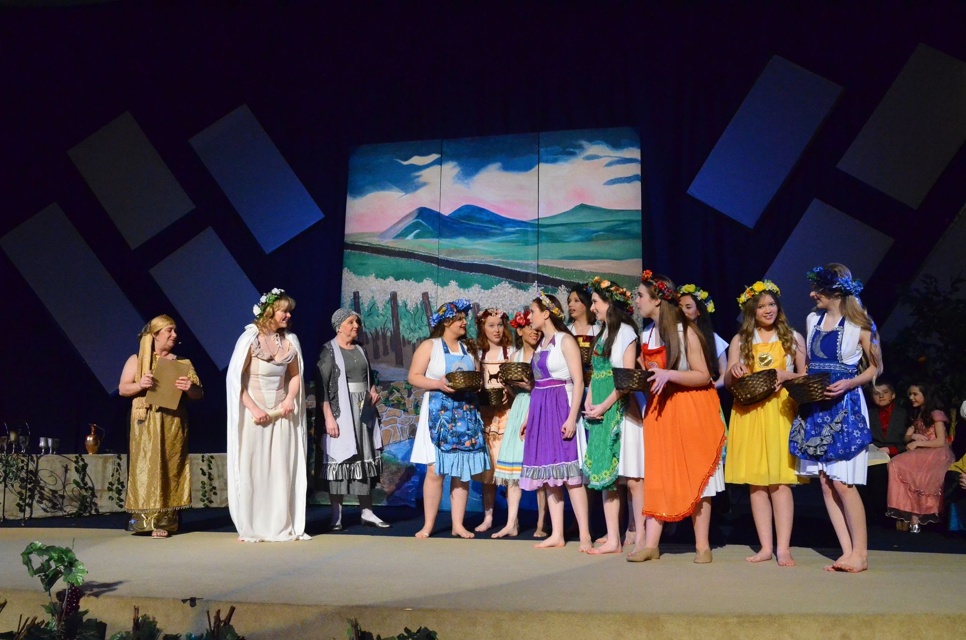 A diverse cast of performers wearing colorful costumes and flower crowns stands on stage before a backdrop depicting a mountainous landscape, with geometric blue shapes flanking the scenic design.