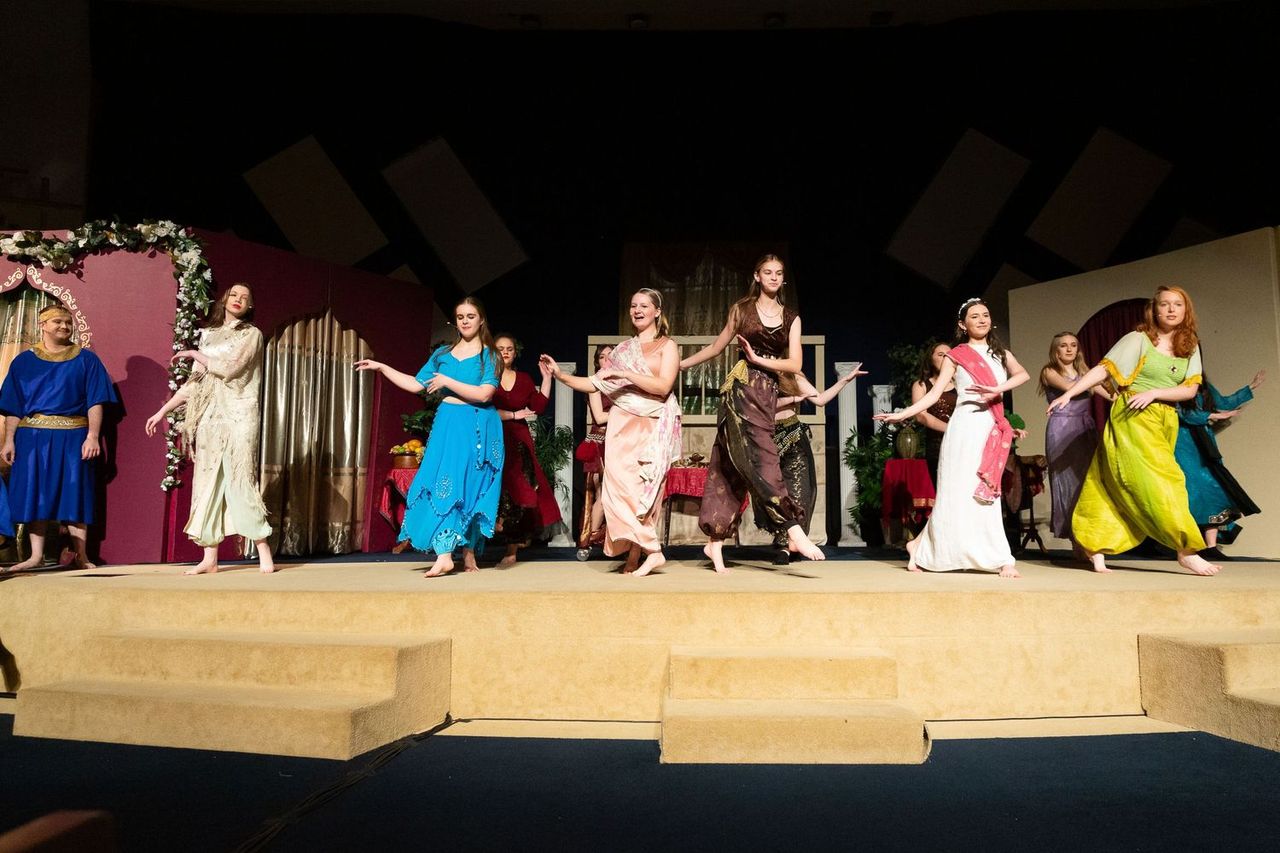 A cast of performers in colorful draped costumes performs on a wooden stage set with classical columns and festive garland decorations against a burgundy and cream backdrop.