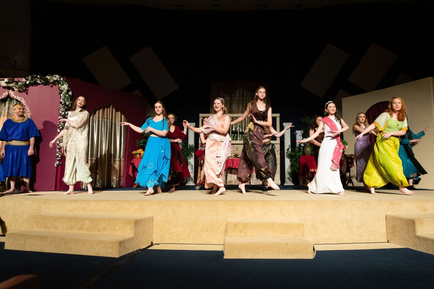 A cast of performers in colorful draped costumes performs on a wooden stage set with classical columns and festive garland decorations against a burgundy and cream backdrop.