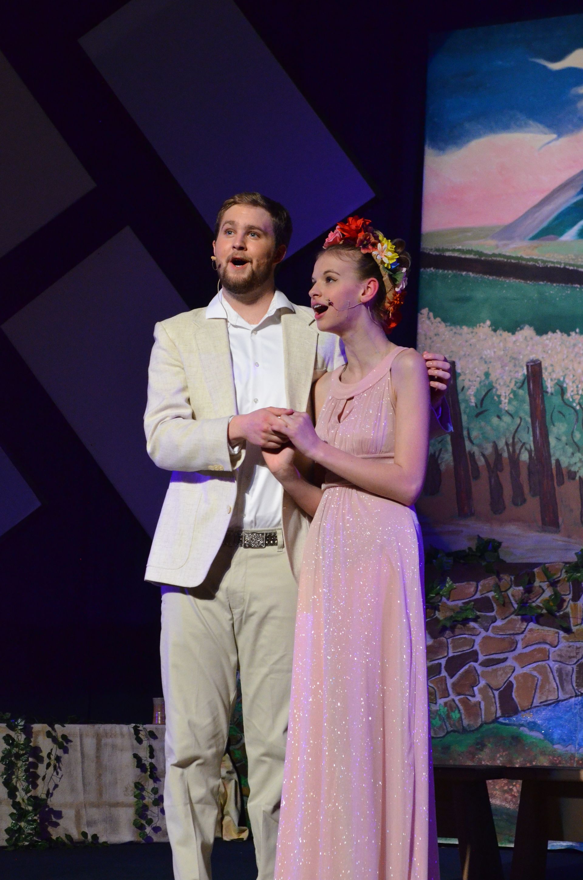 Two performers in cream-colored costumes stand together on a dark stage with colorful projected scenery behind them, one wearing a floral crown and pink shawl.