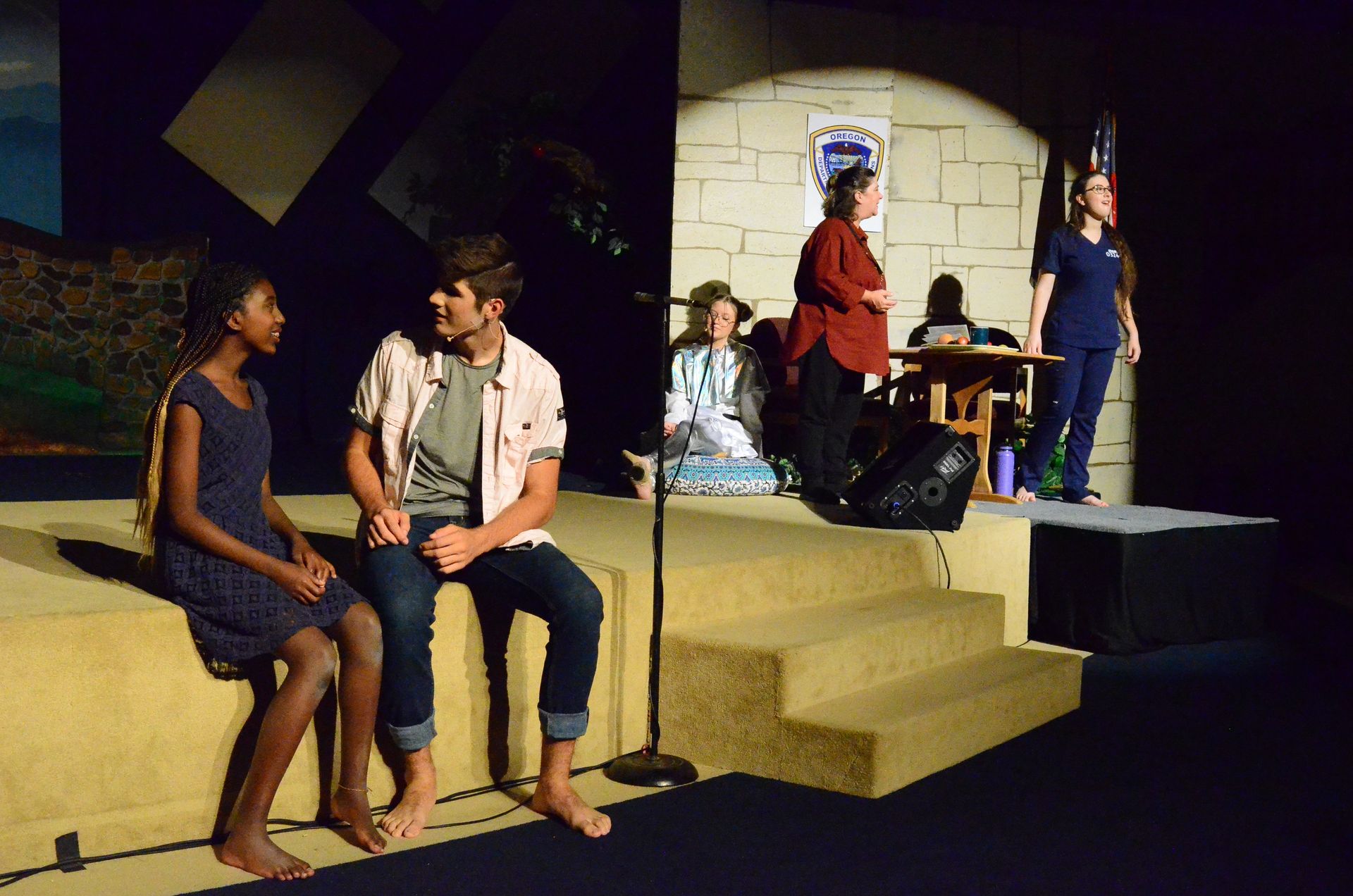 Two performers sit on a yellow platform stage engaging in conversation while other cast members stand and sit in the background of a stone-walled set under theatrical lighting.