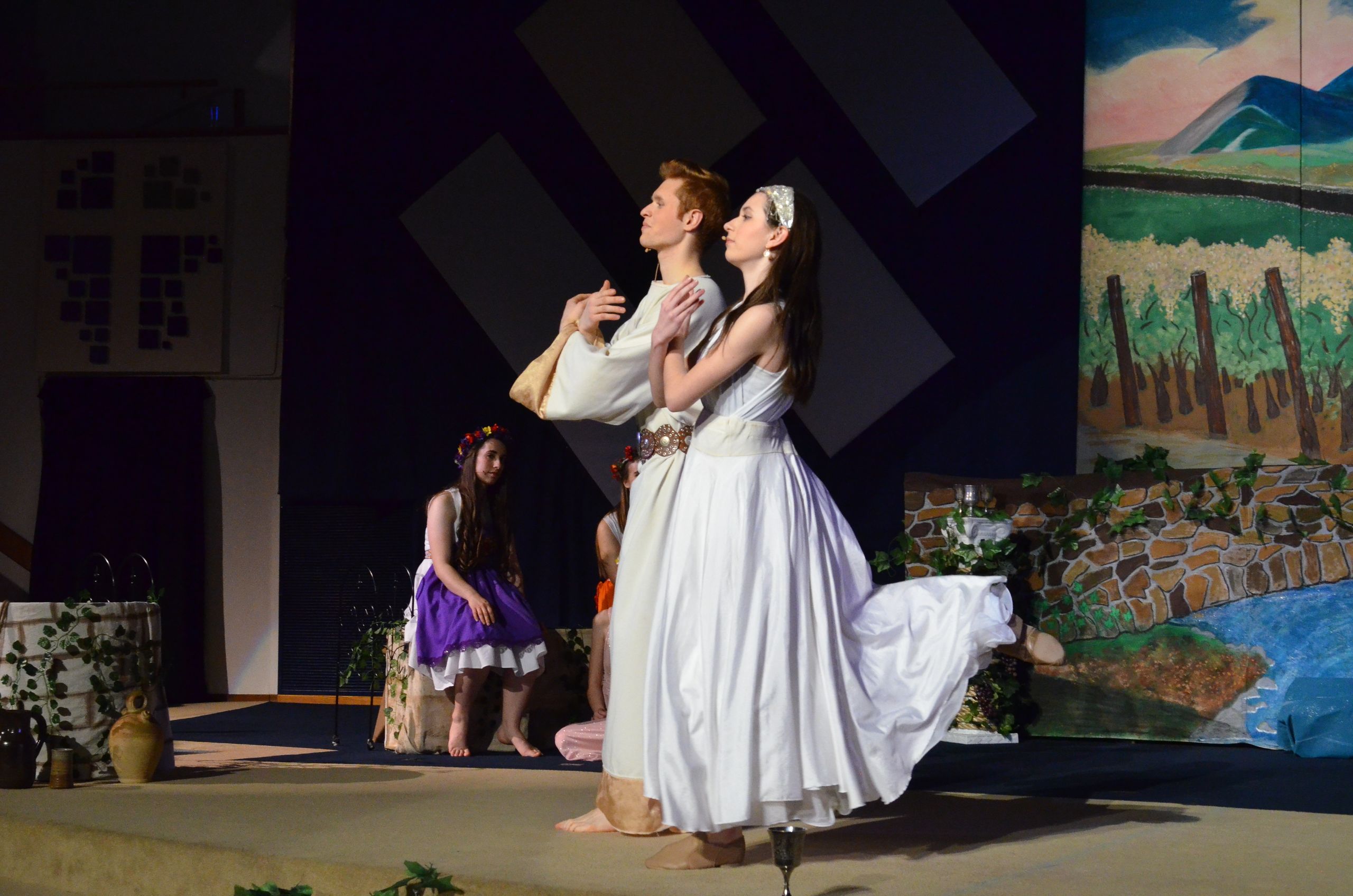 Two performers in white flowing garments stand center stage gazing upward, with a smaller performer in a purple dress beside them, set against geometric backdrop panels and a painted landscape scenery depicting fields and sky.