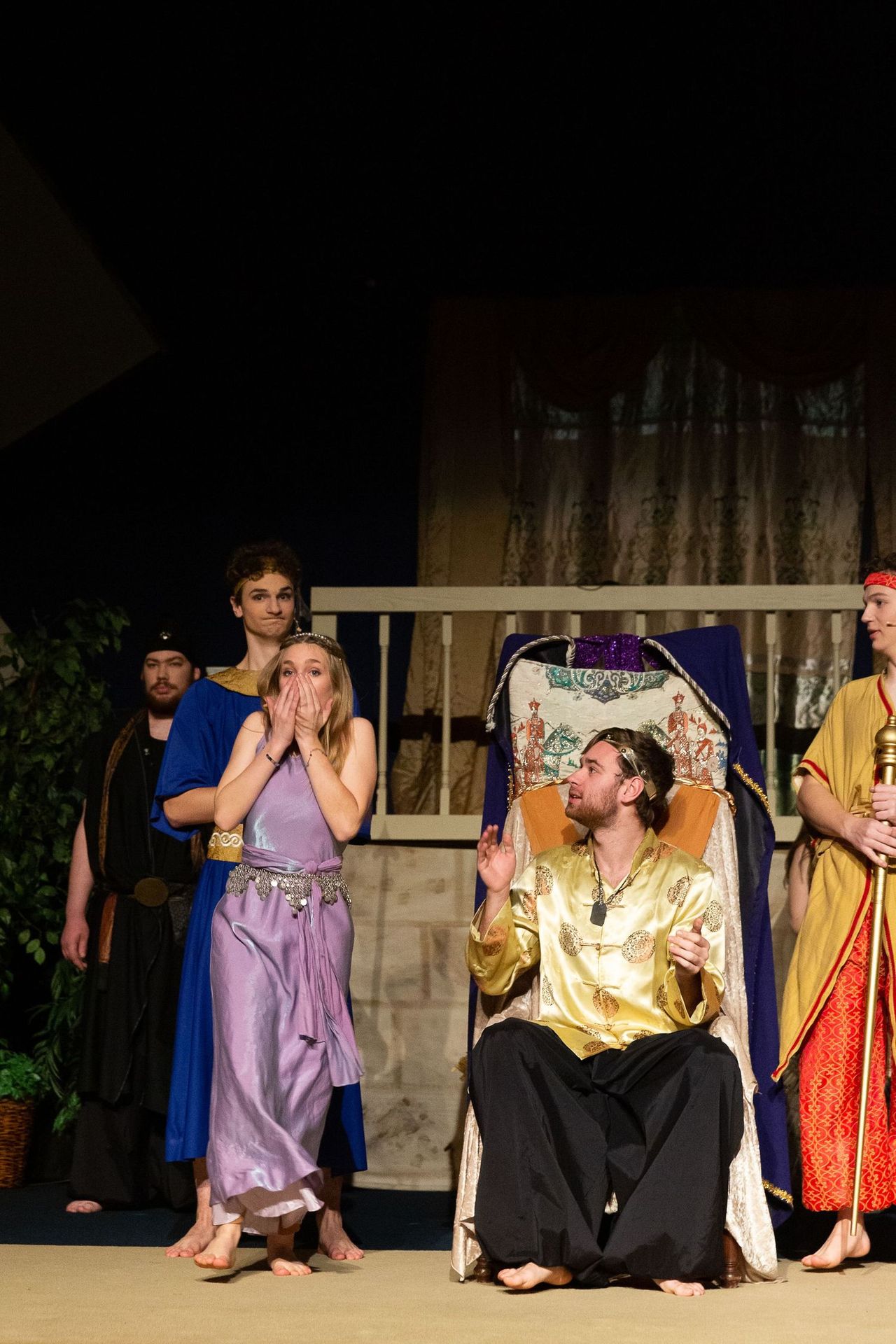 A cast of performers in colorful robes and tunics stand on stage around a seated performer in a golden garment, with one performer holding their hands to their face in an expression of concern or surprise.