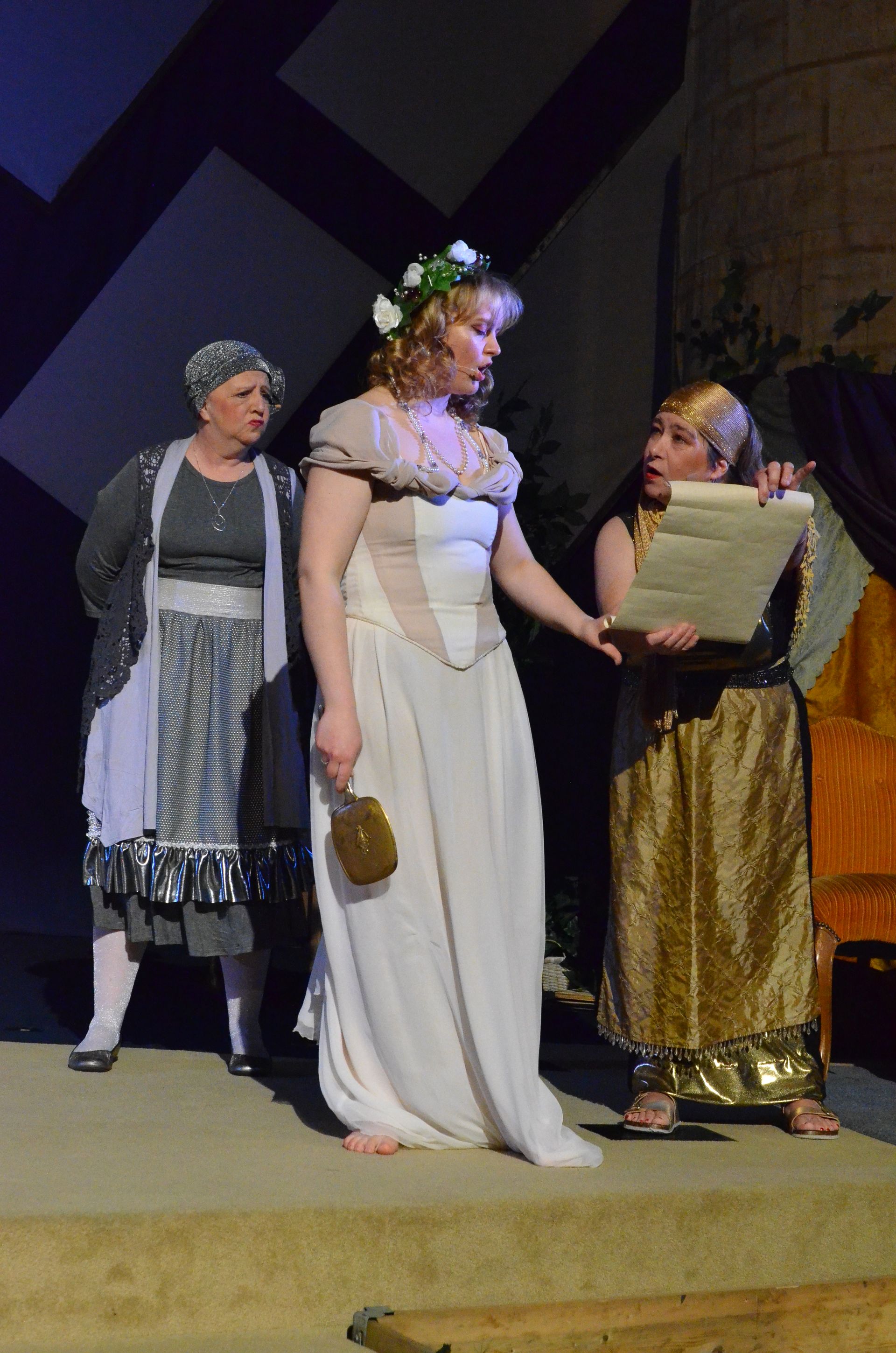 Two performers on stage, one seated in a white costume holding a gold prop while reading from a script, and another standing beside them in period costume against a golden backdrop.