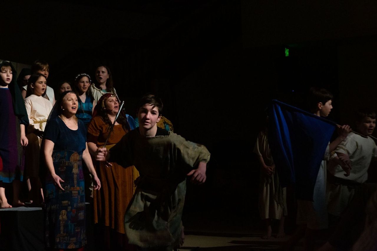 A cast of performers in period costumes stands on a dimly lit stage, with some looking upward in wonder as others hold props and fabrics, creating a dramatic scene lit by theatrical spotlights against a dark background.