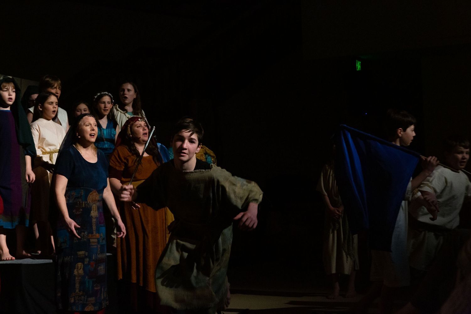 A cast of performers in period costumes stands on a dimly lit stage, with some looking upward in wonder as others hold props and fabrics, creating a dramatic scene lit by theatrical spotlights against a dark background.