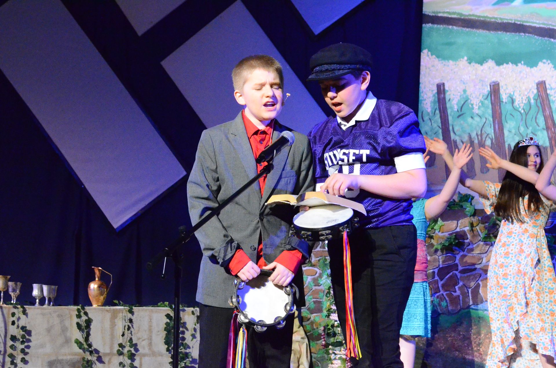 Two performers on stage hold a tambourine-like prop while singing, with one wearing a grey blazer and red bow tie and the other wearing a blue cap and blue shirt, surrounded by cast members in colorful costumes against a painted backdrop depicting a beach scene with wooden posts.