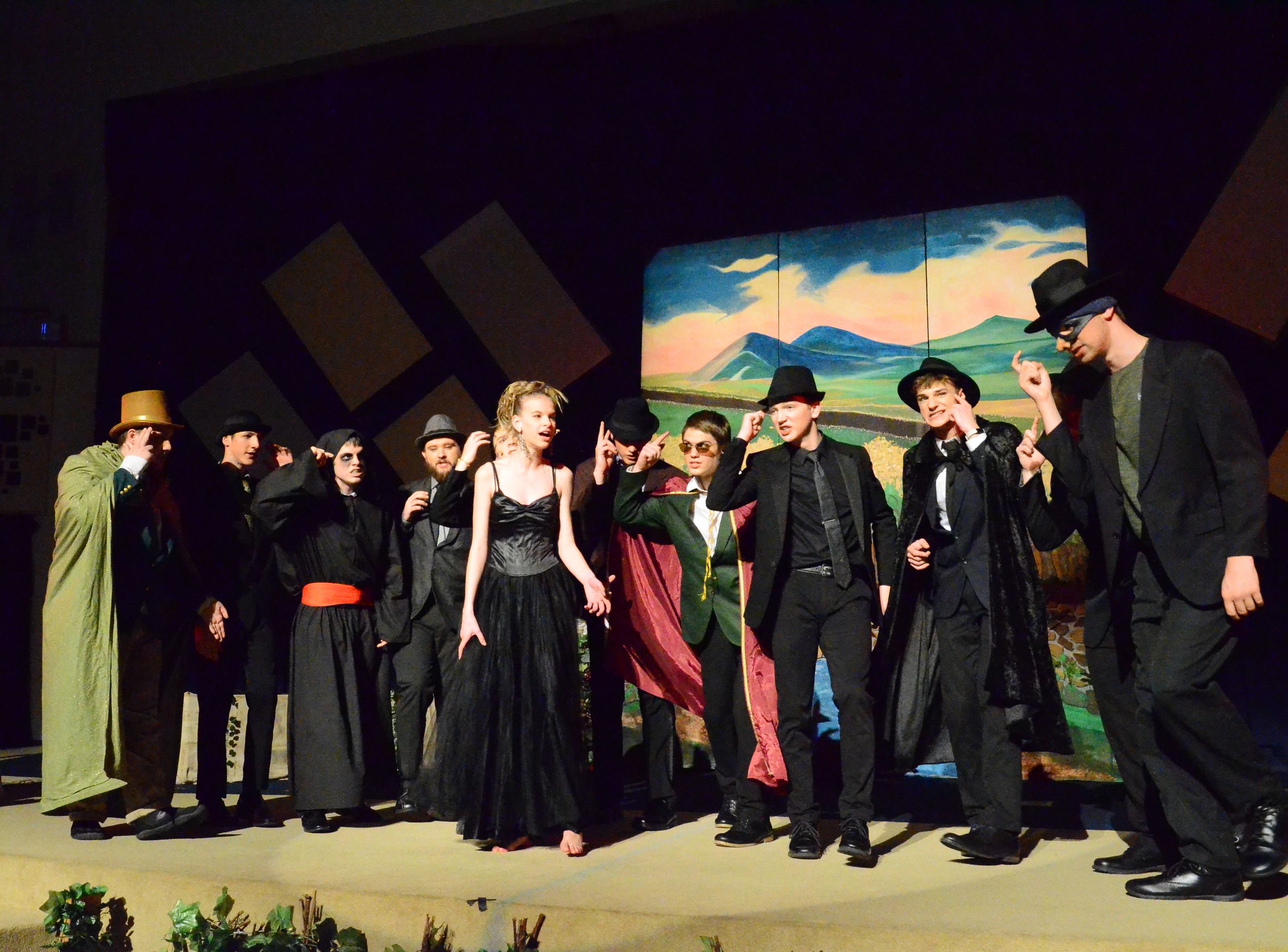 A cast of performers in dark formal attire and hats stands on stage before a painted backdrop depicting a pastoral mountain landscape, with some cast members gesturing expressively during what appears to be a musical theatre production.