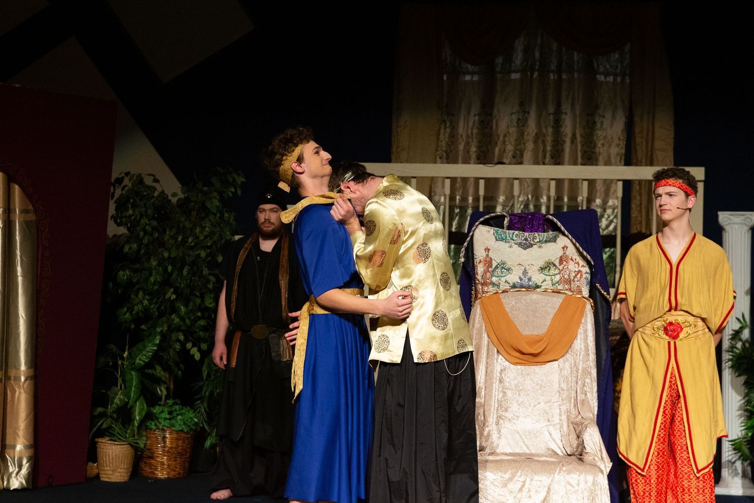 A cast of performers in colorful biblical-style robes and headpieces stand on stage among potted plants and draped fabrics, with one performer in a blue robe and gold sash gesturing expressively while others look on.