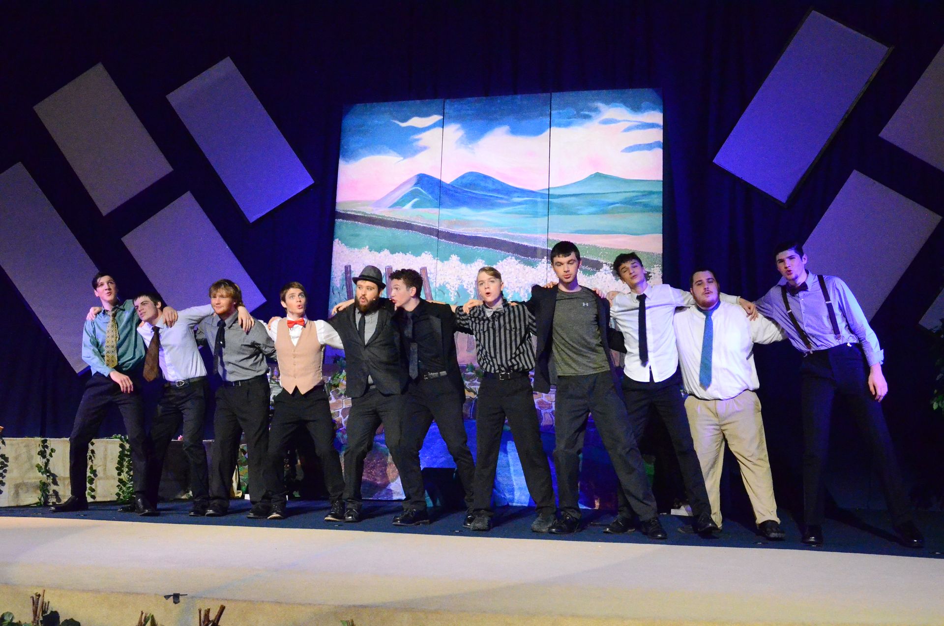 A cast of ten performers stands in a line across a stage wearing formal attire, with their arms around each other's shoulders, positioned in front of a large backdrop displaying a scenic landscape with mountains and water.